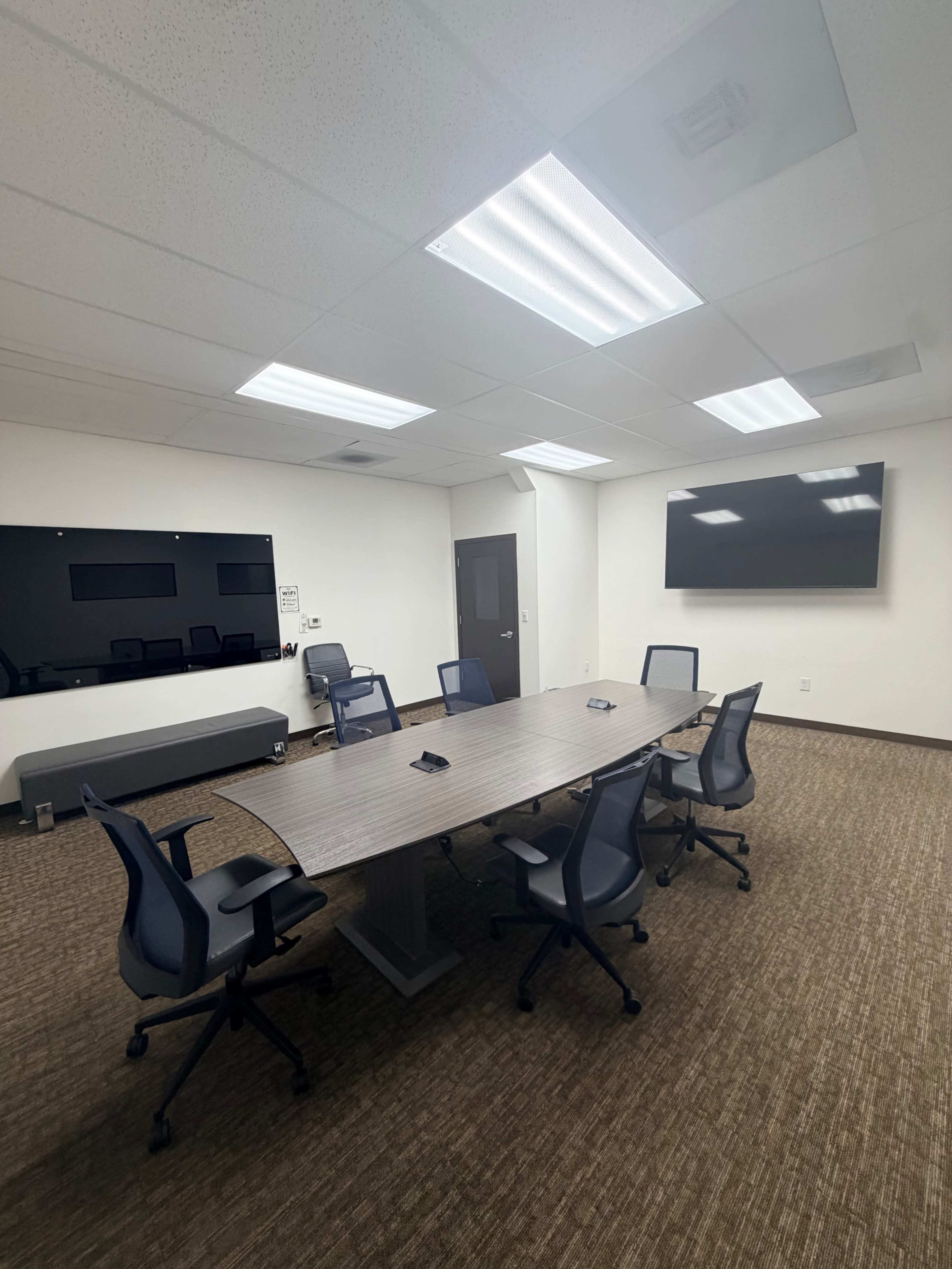 The image shows a modern conference room with a large table surrounded by six chairs, two wall-mounted screens, and bright overhead lighting.