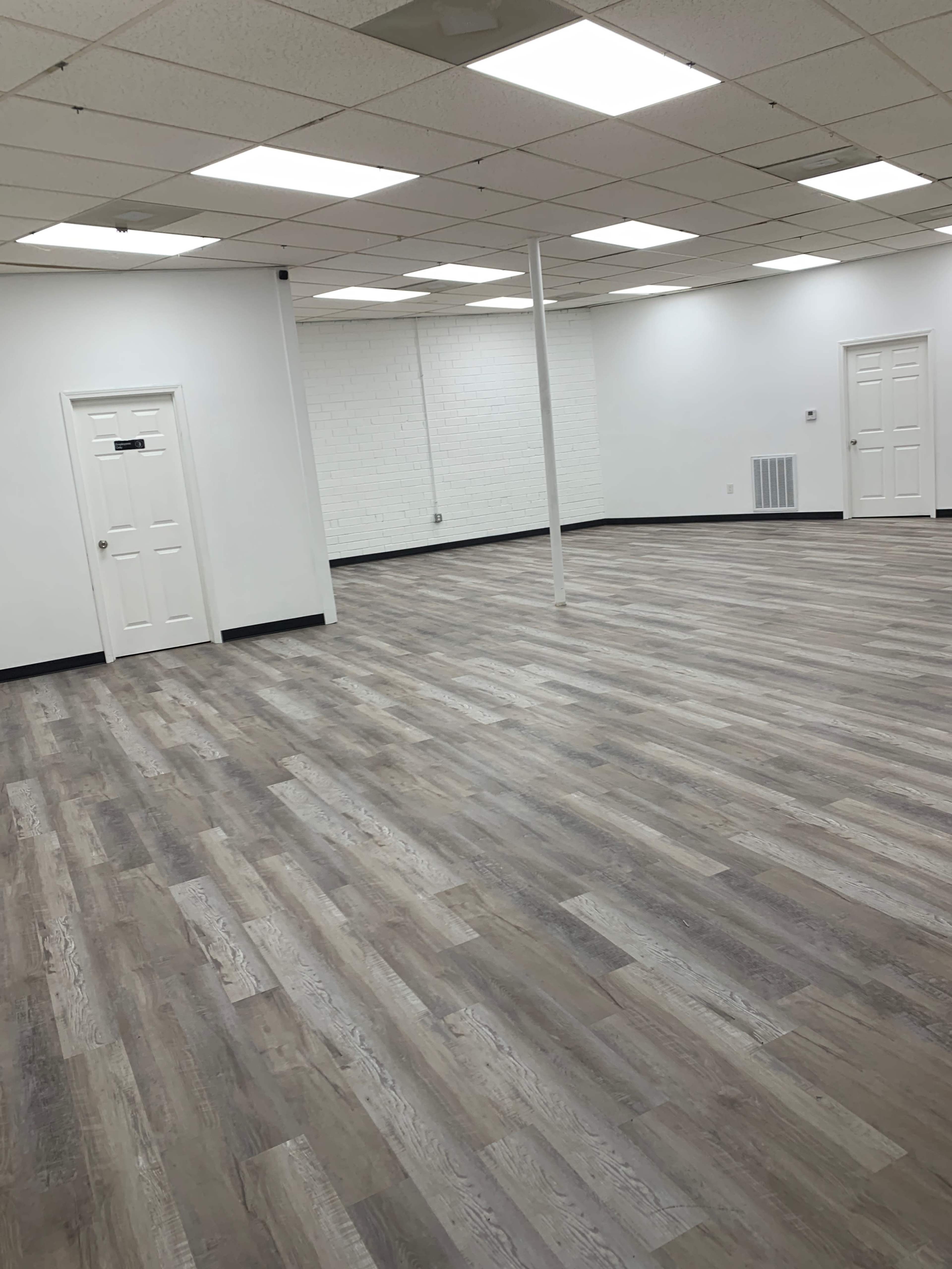 The image shows a spacious, empty room with wooden floors, white walls, and two doors.