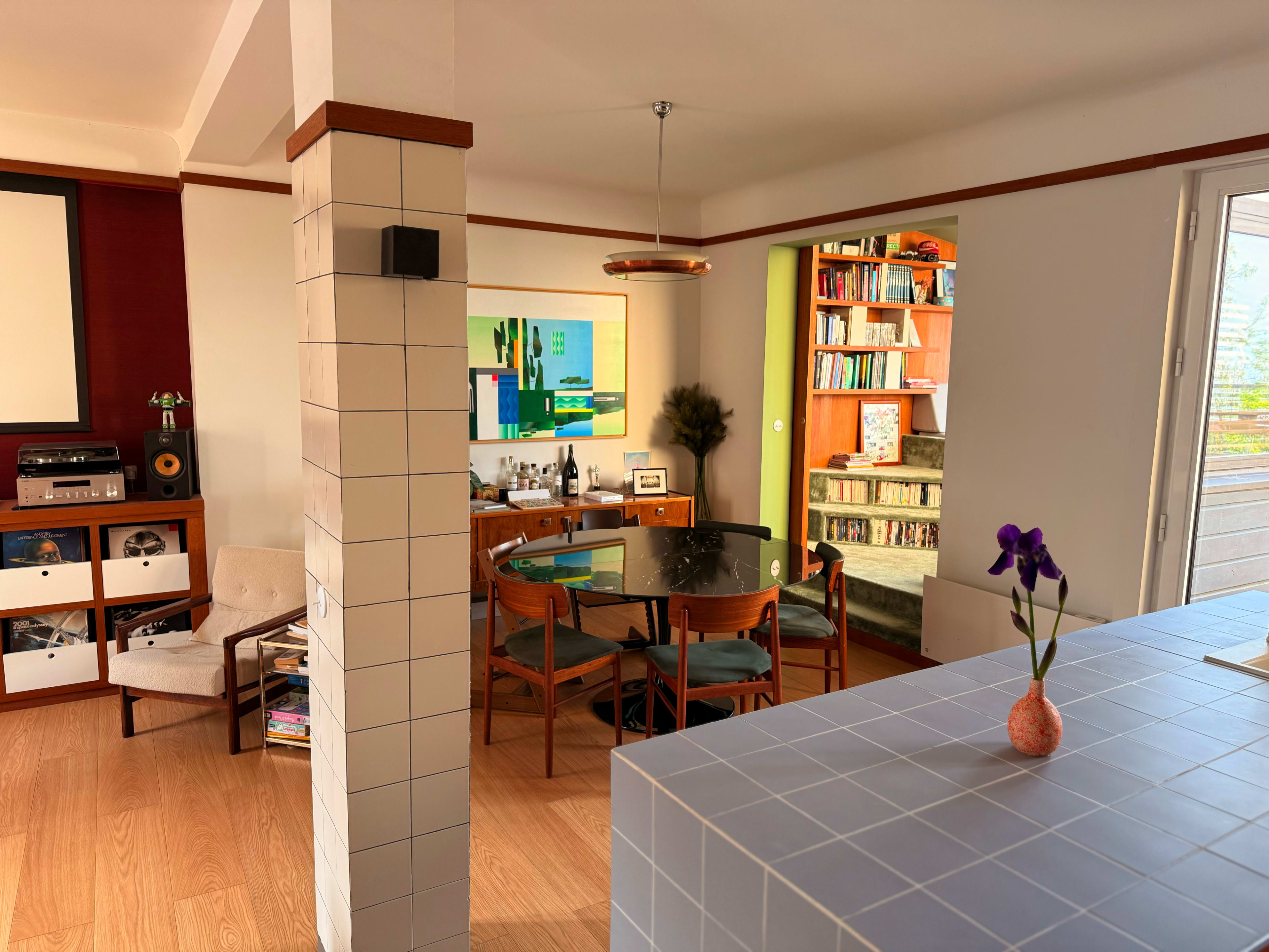 The image shows a modern living space featuring a dining area with a round glass table, chairs, and a bookshelf in the background, along with a kitchen counter with a small flower vase.