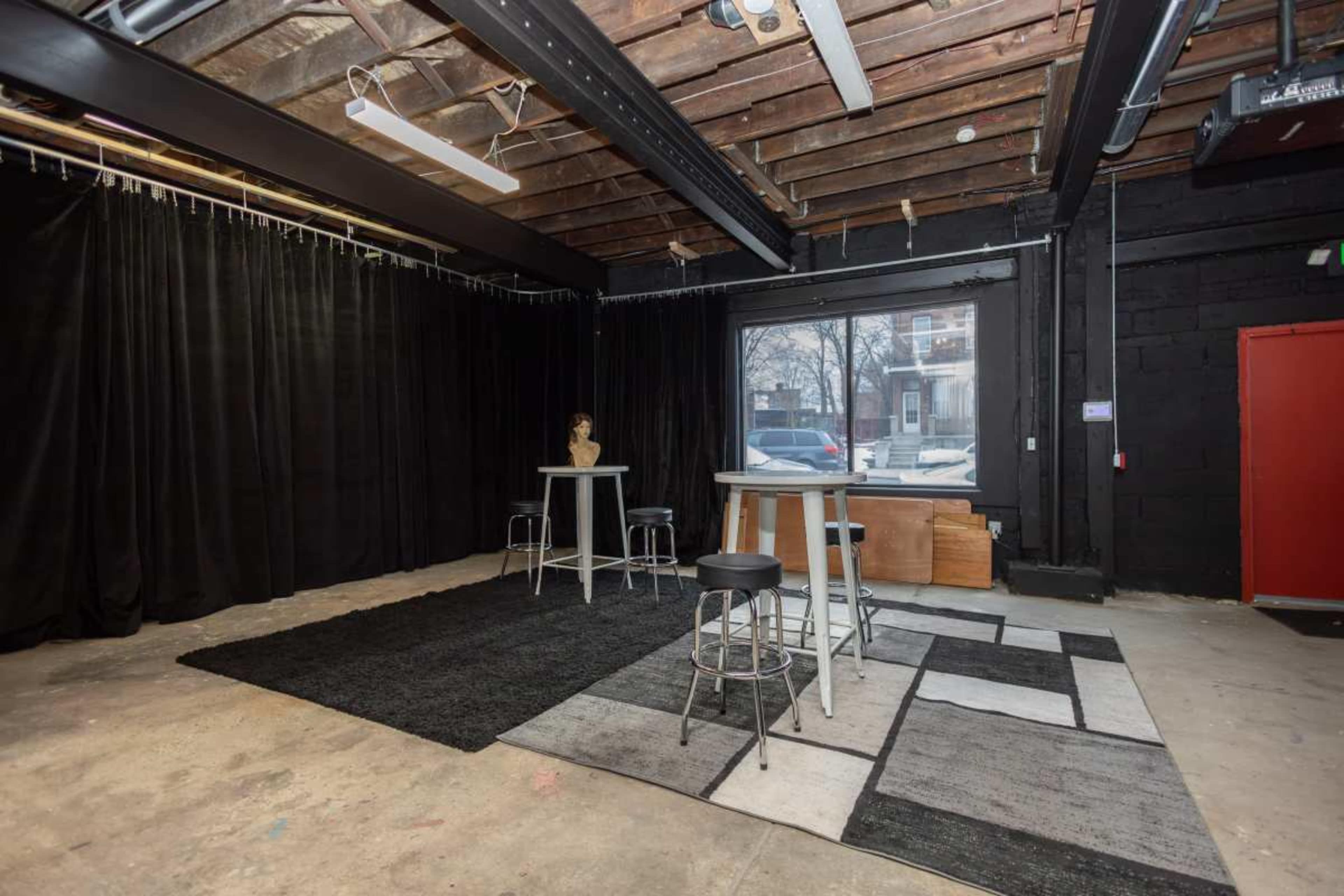 The image shows an empty interior space with a black backdrop, three tall tables, two stools, and a window overlooking a parking area.