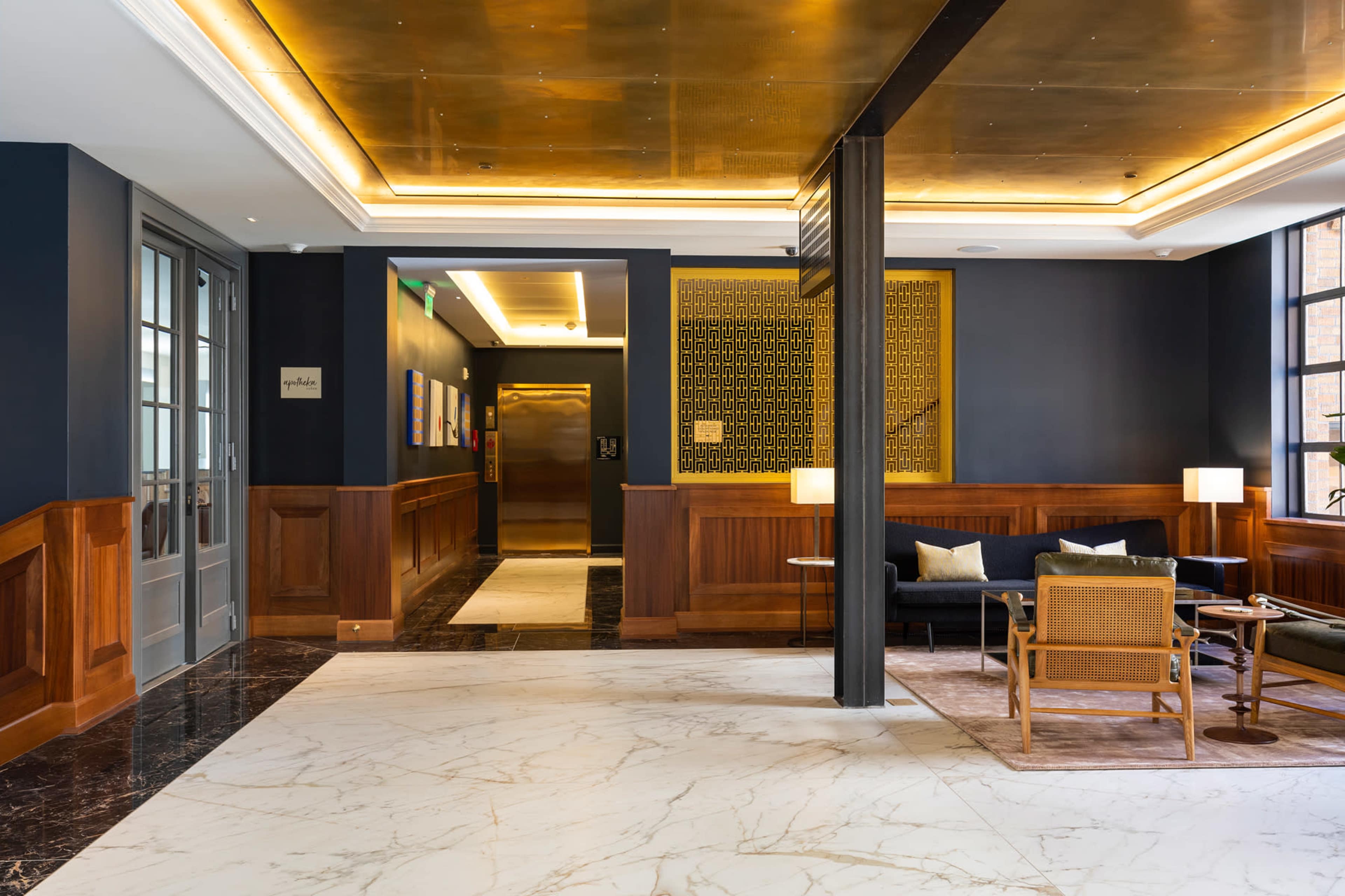 A modern hotel lobby featuring wood paneling, marble flooring, and seating arrangements near a glass door and elevator.