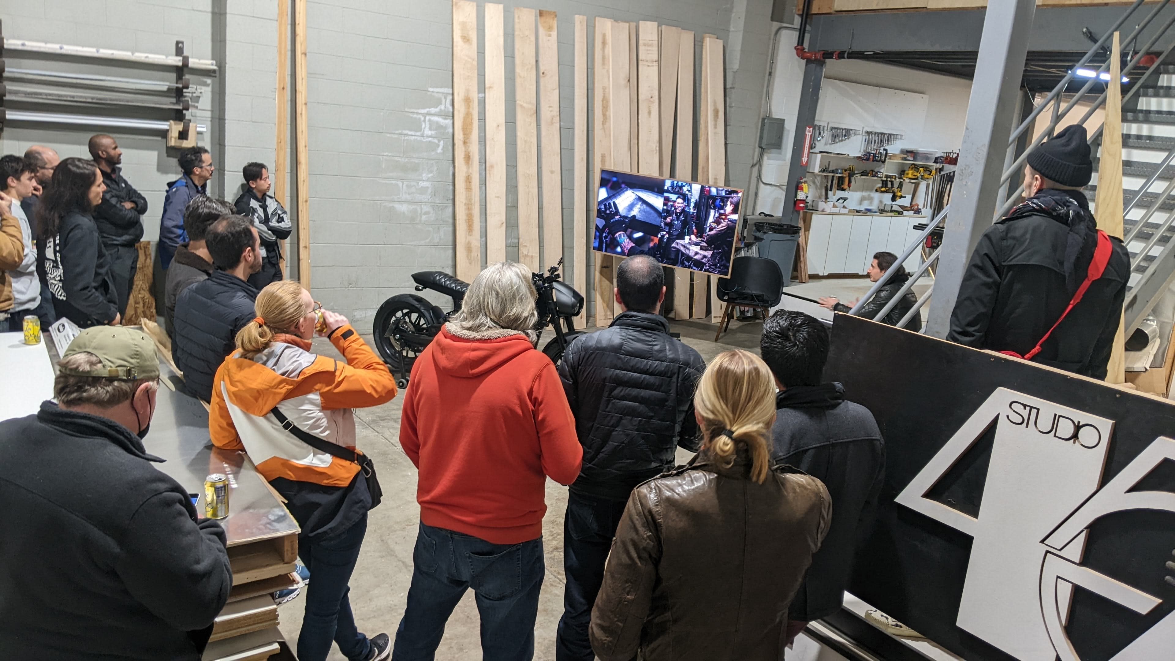 A group of people is gathered in a workshop, watching a presentation on a large screen while surrounded by various tools and equipment.