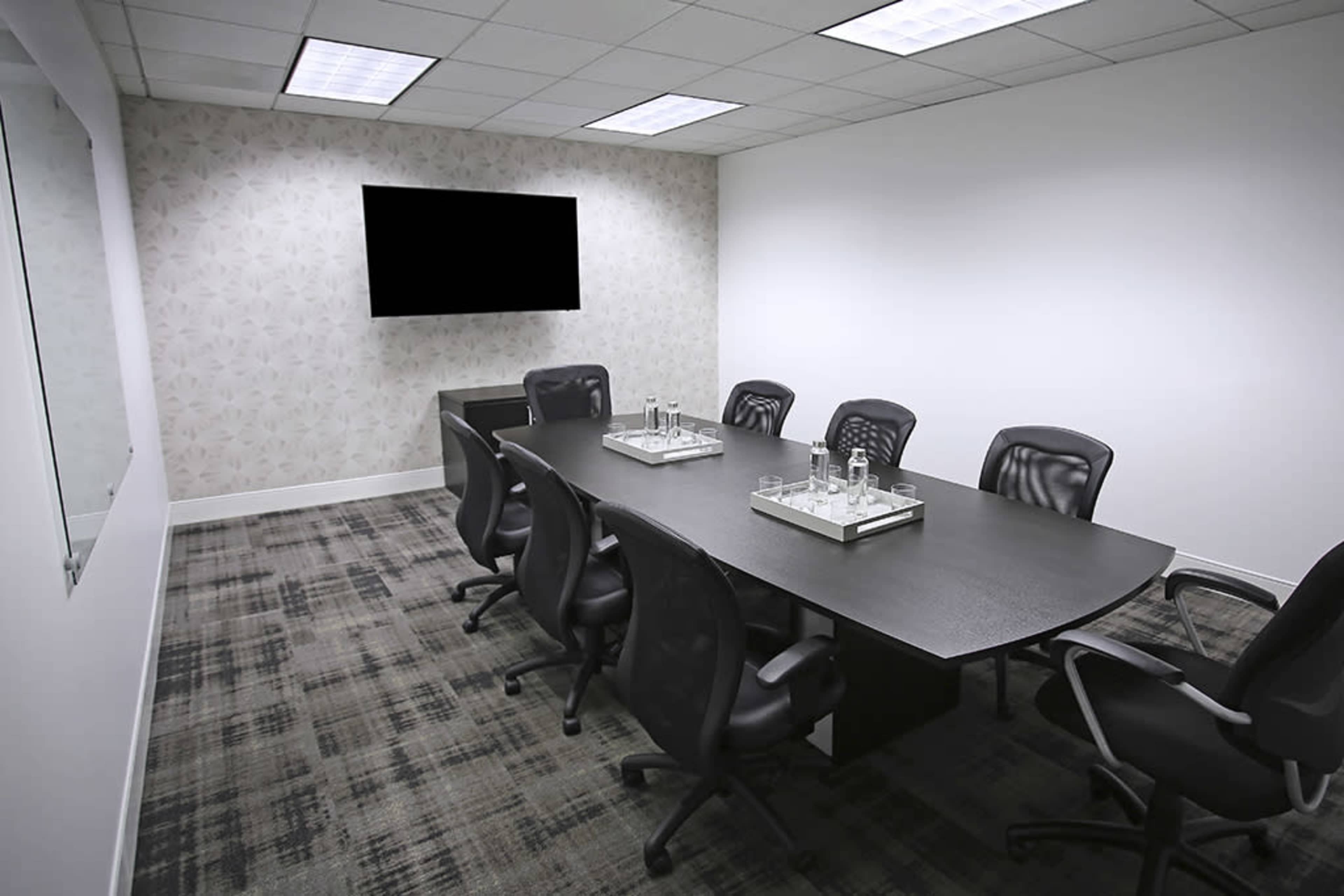 A modern conference room with a large table surrounded by ergonomic chairs, a television mounted on the wall, and a patterned backdrop.