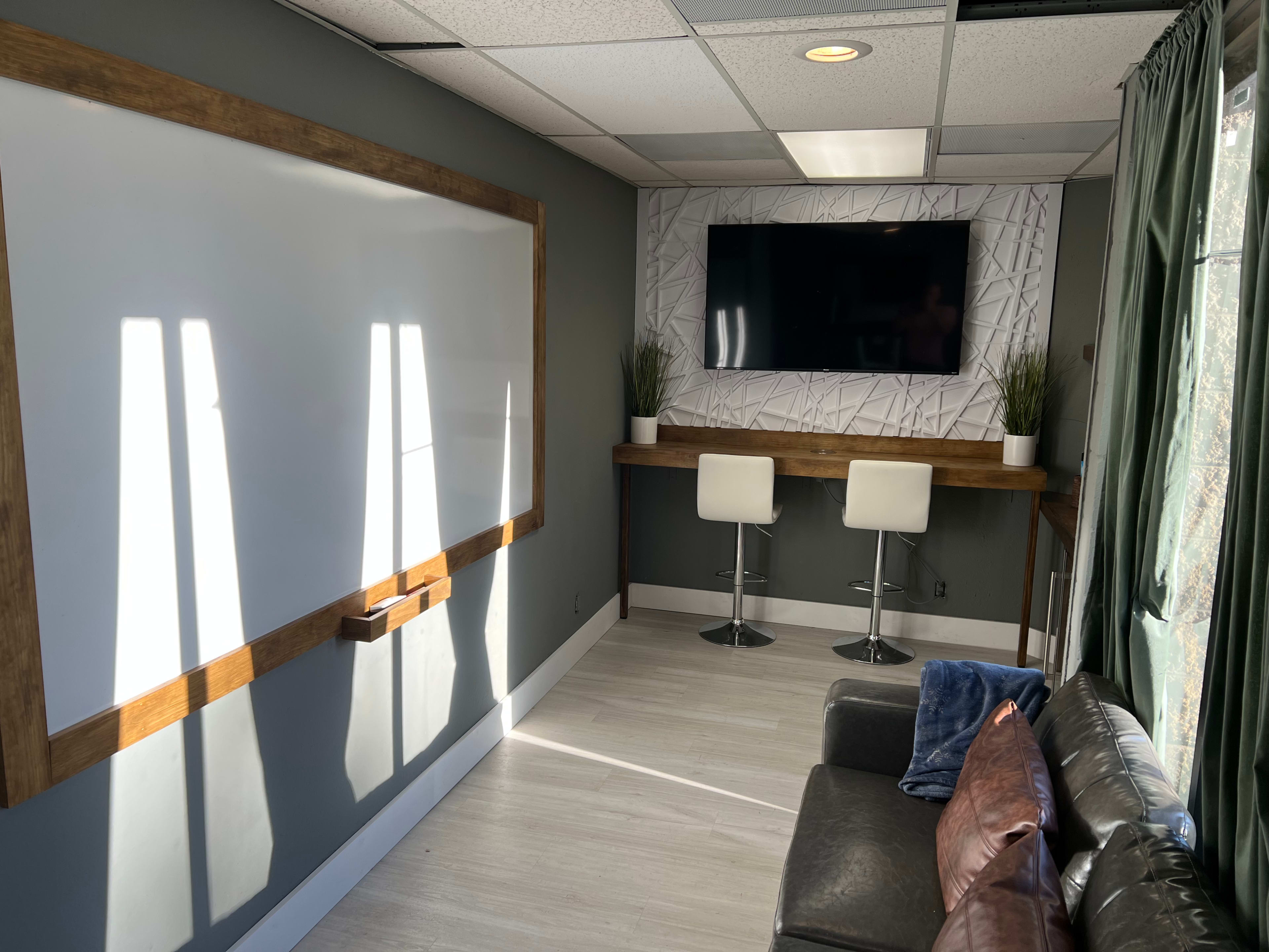 A small meeting room with a whiteboard, a TV mounted on the wall, a wooden counter with two white stools, and a couch in the corner.