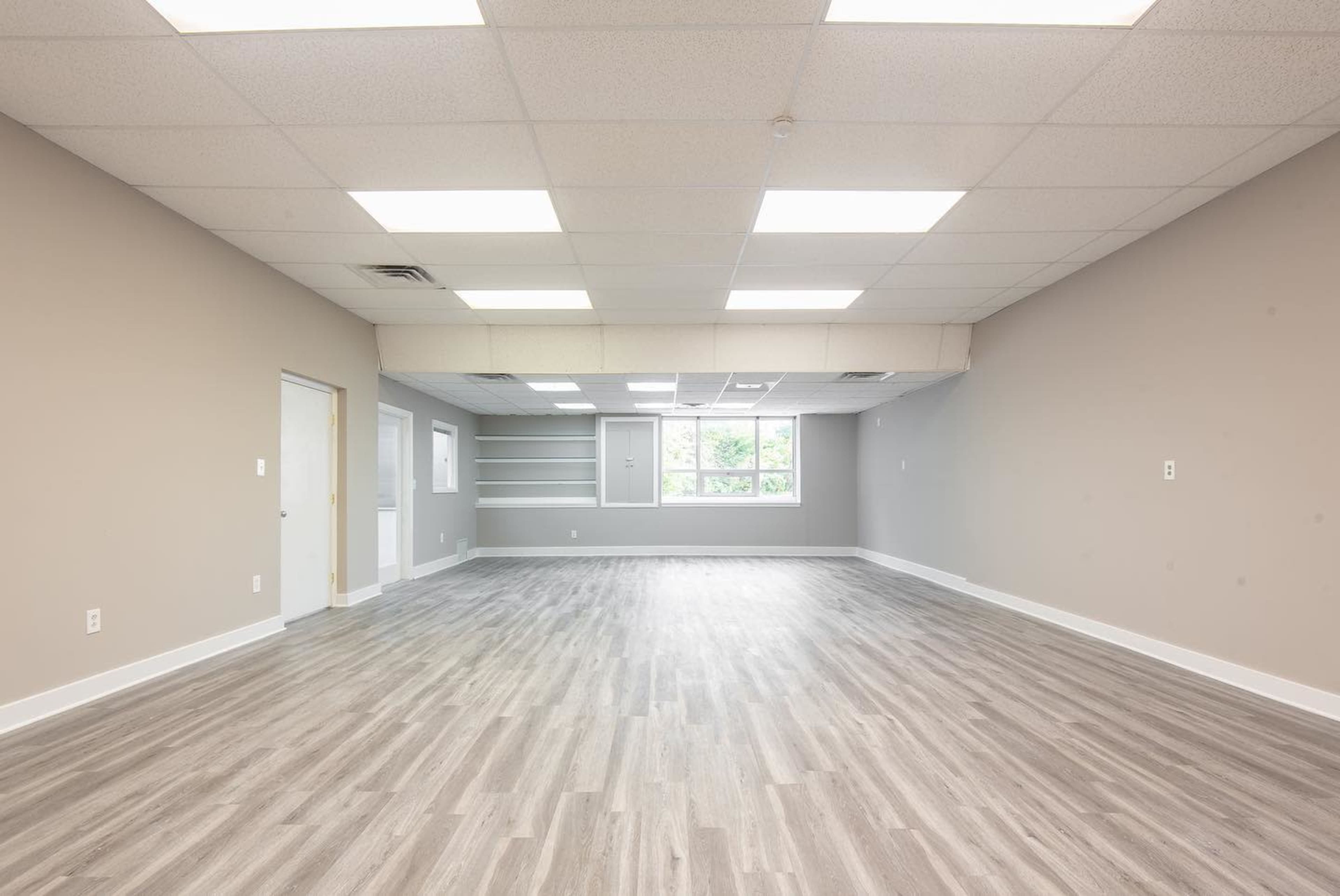 The image shows a large, empty room with gray walls, a light wooden floor, and several ceiling lights.
