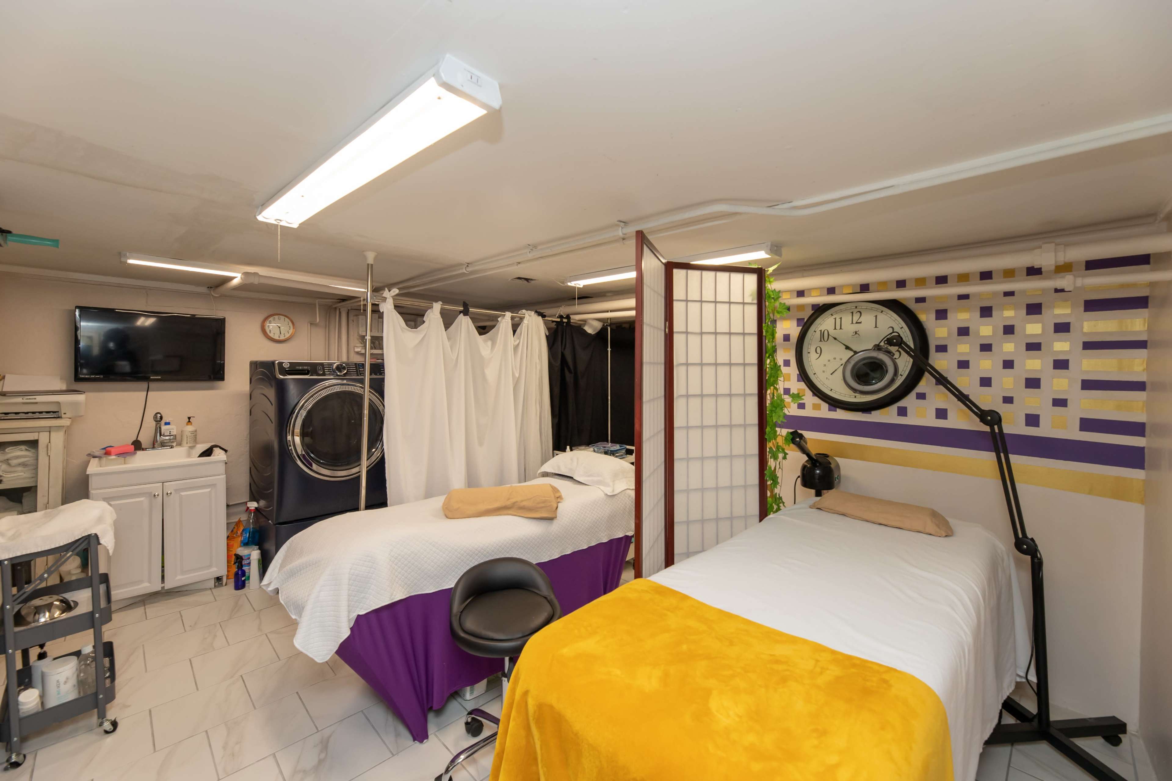 The image shows a treatment room in a basement featuring two massage tables, a washing machine, and various treatment equipment.