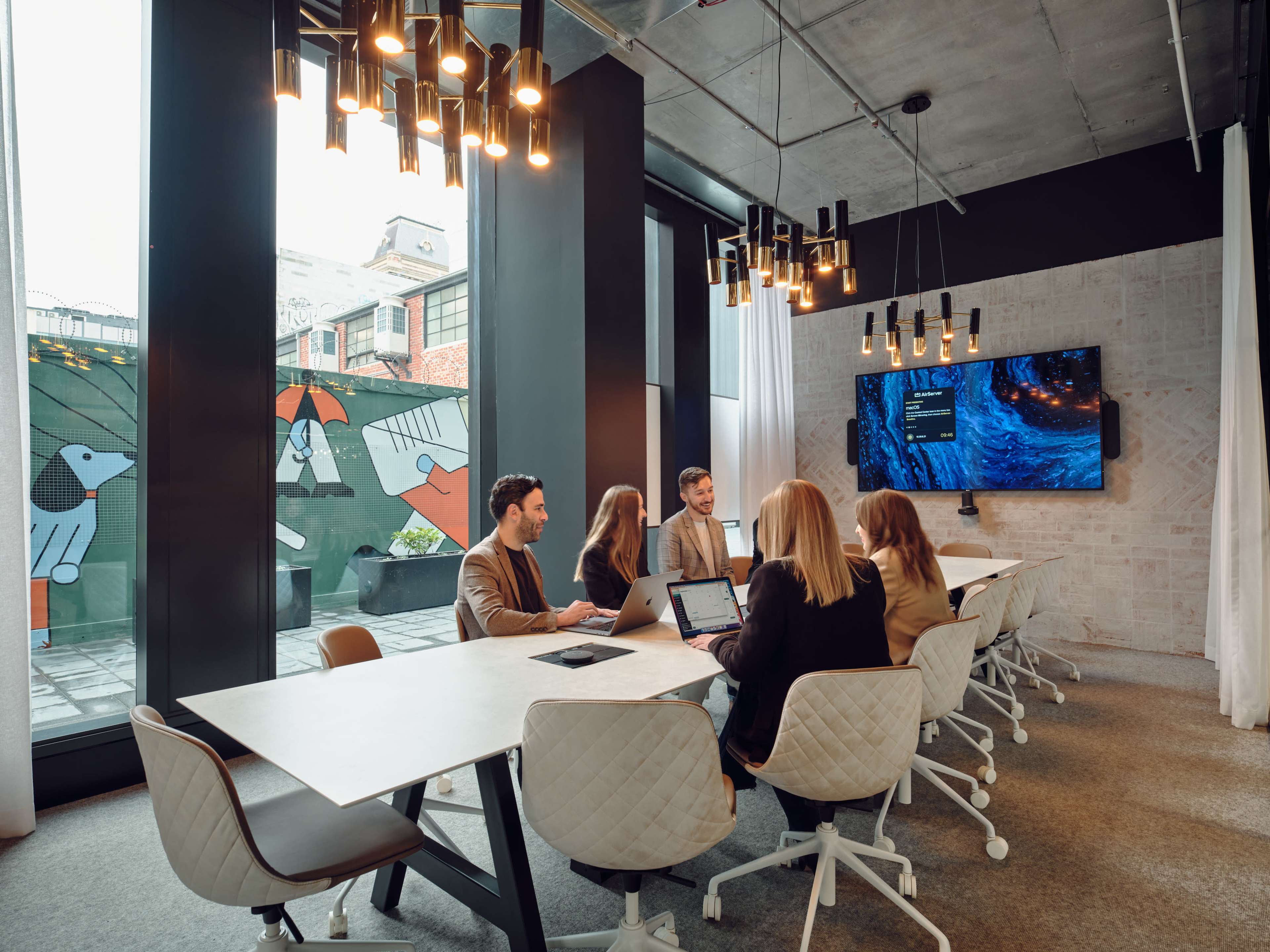 A group of five people are seated around a conference table in a modern meeting room, with large windows displaying a colorful mural outside.