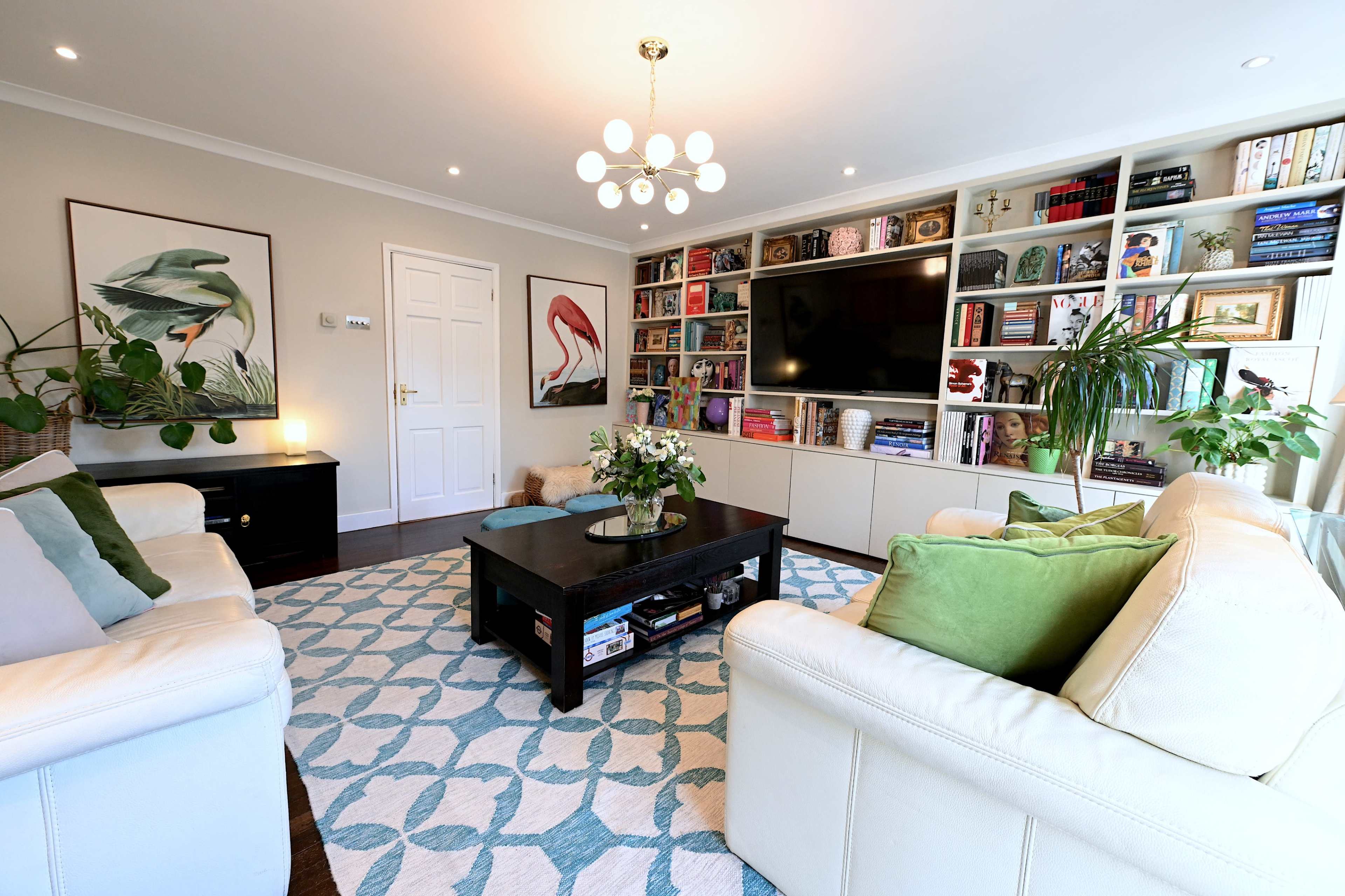 The image shows a modern living room featuring two white sofas, a dark coffee table, and shelves filled with books and decor, illuminated by a decorative ceiling light.