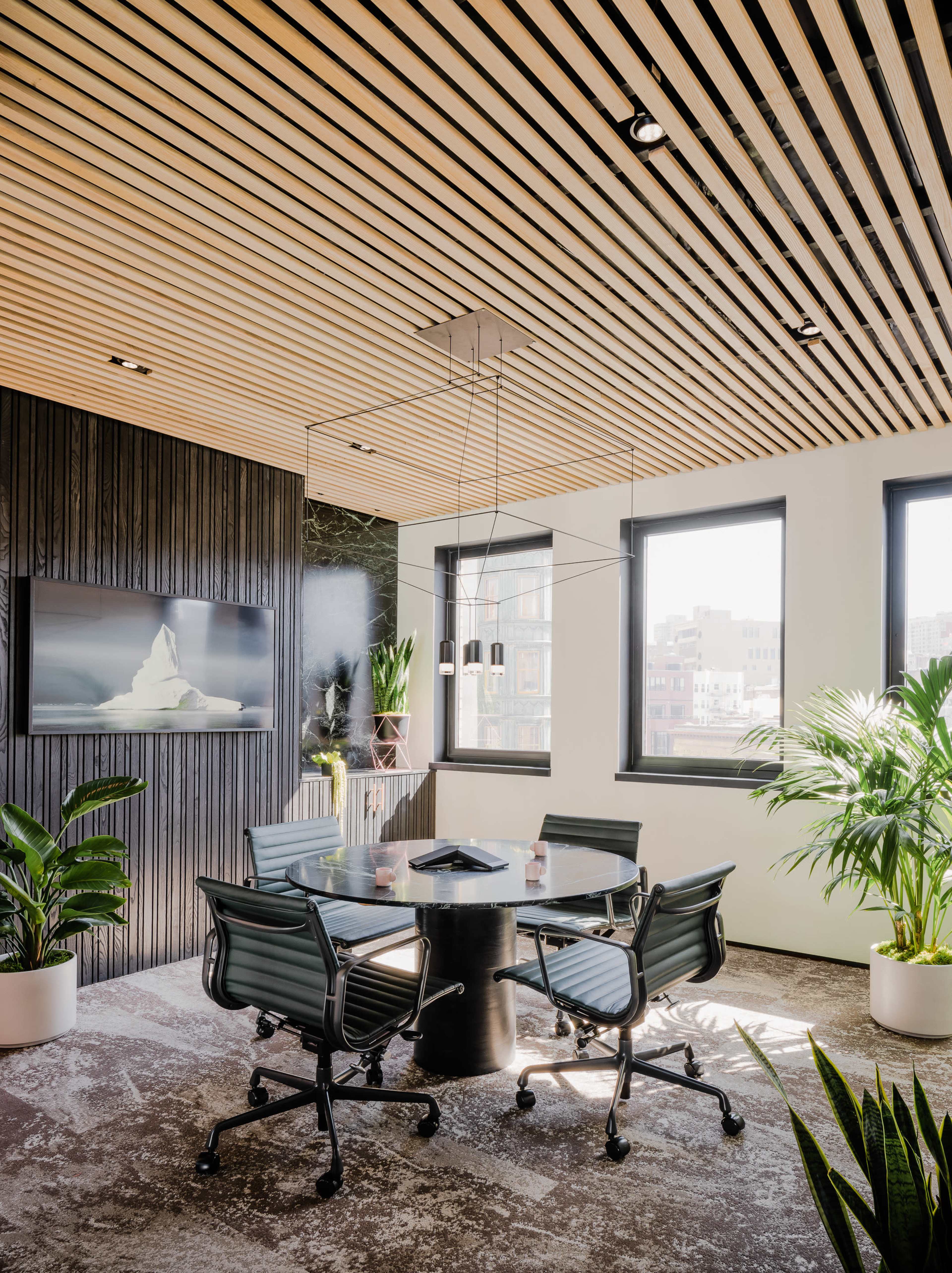 The image shows a modern conference room with a round table, black office chairs, and large windows featuring greenery and city views.