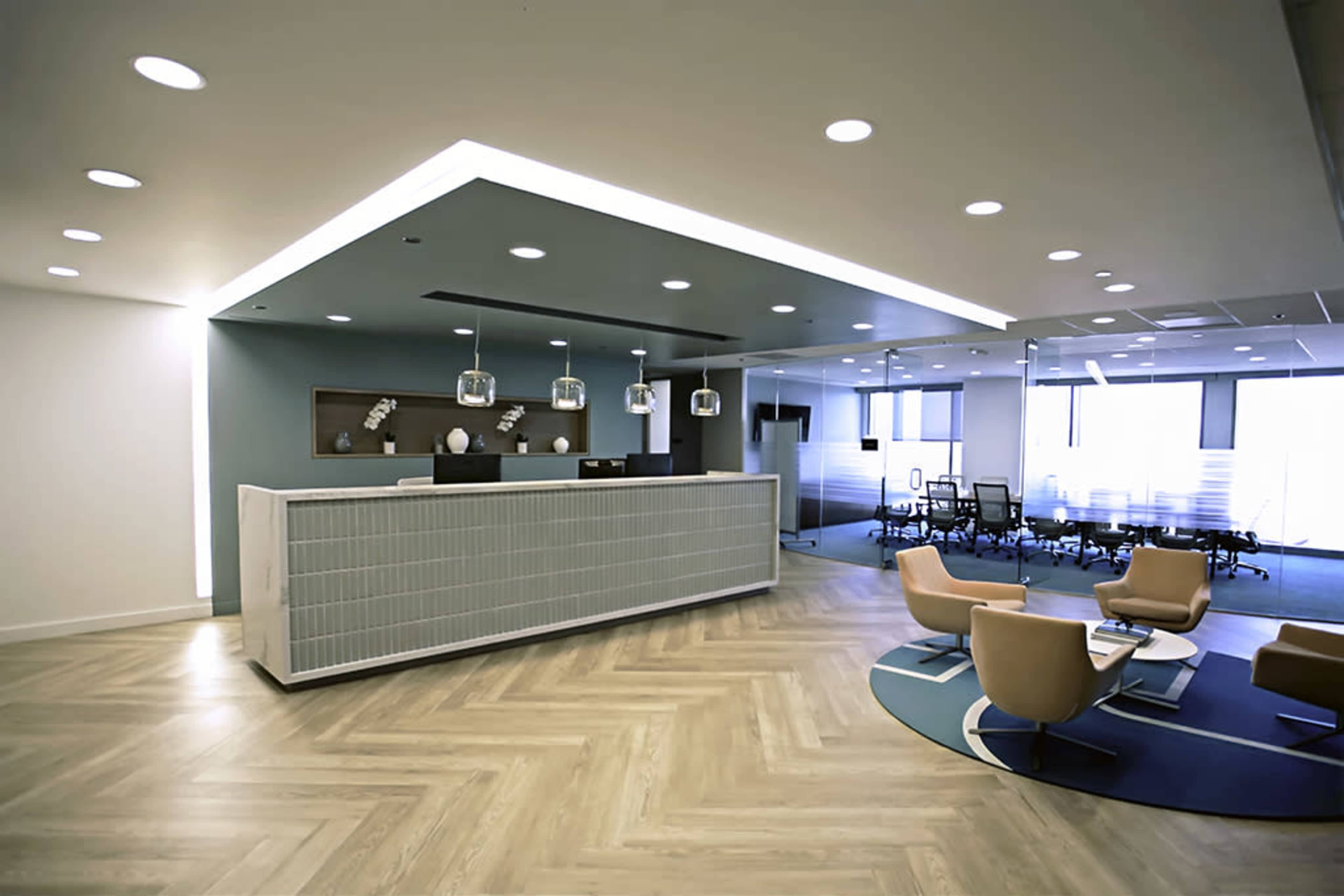A modern office reception area features a sleek front desk, pendant lighting, and seating arrangements with accent chairs and a round rug.
