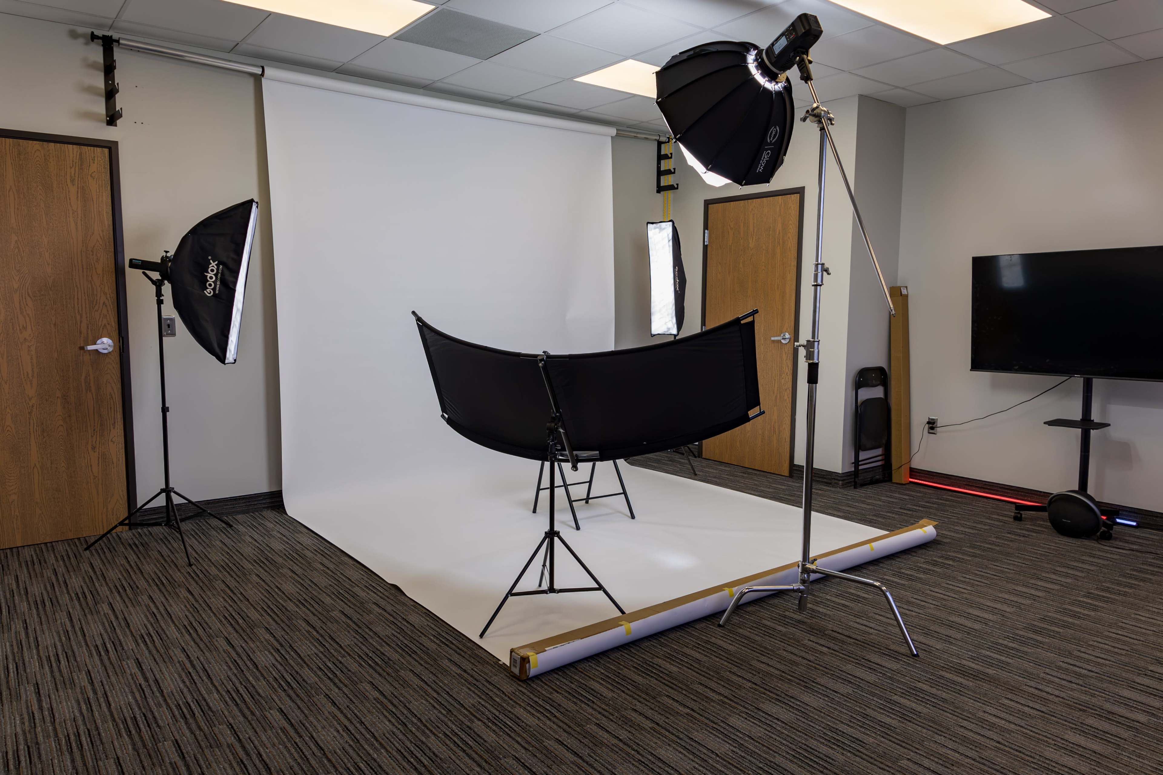 The image shows a photography studio setup with a backdrop, a curved chair, and two softbox lights positioned on either side.