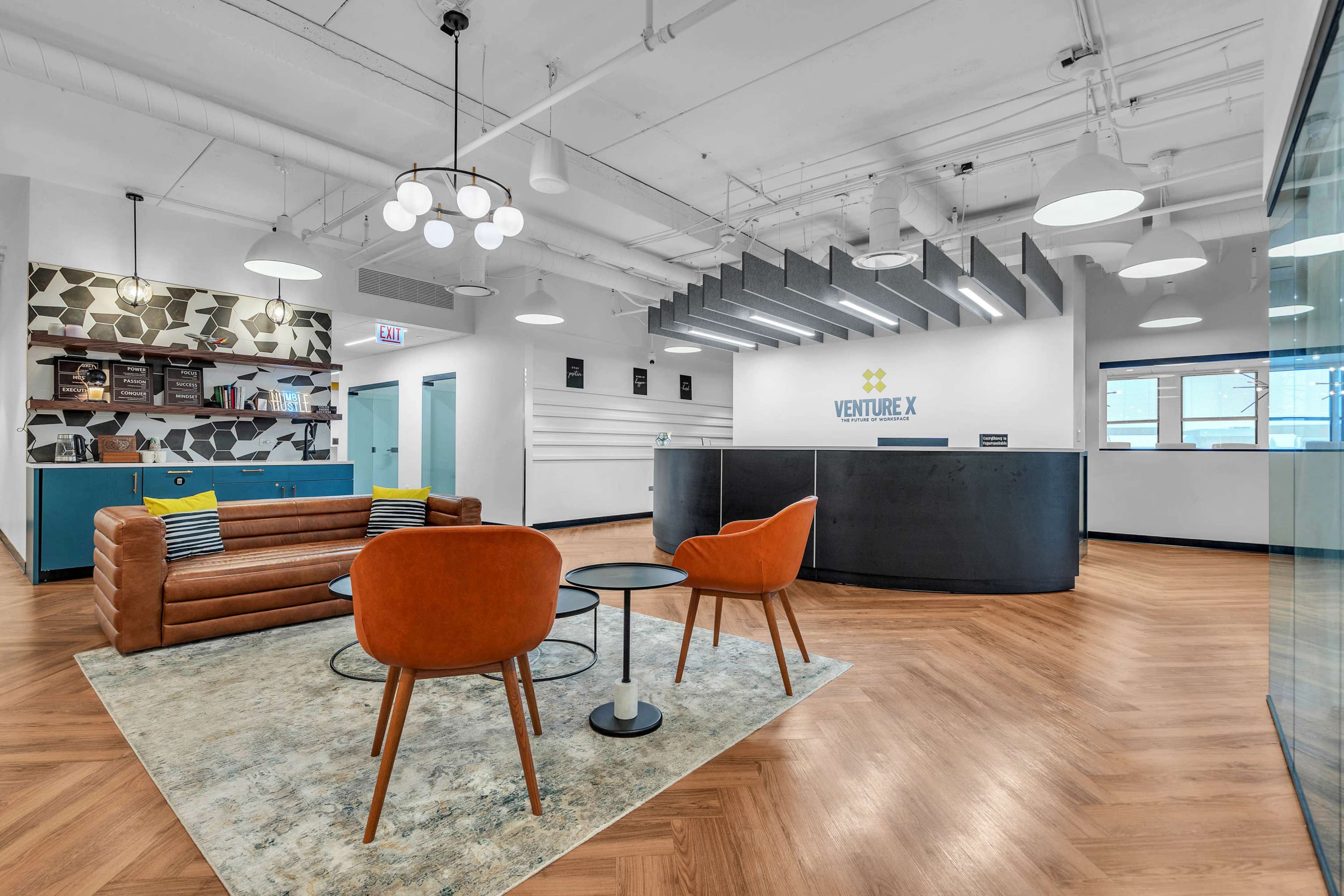 The image shows a modern office reception area with a brown leather sofa, orange chairs, a round table, and a front desk displaying the "VENTURE X" logo.