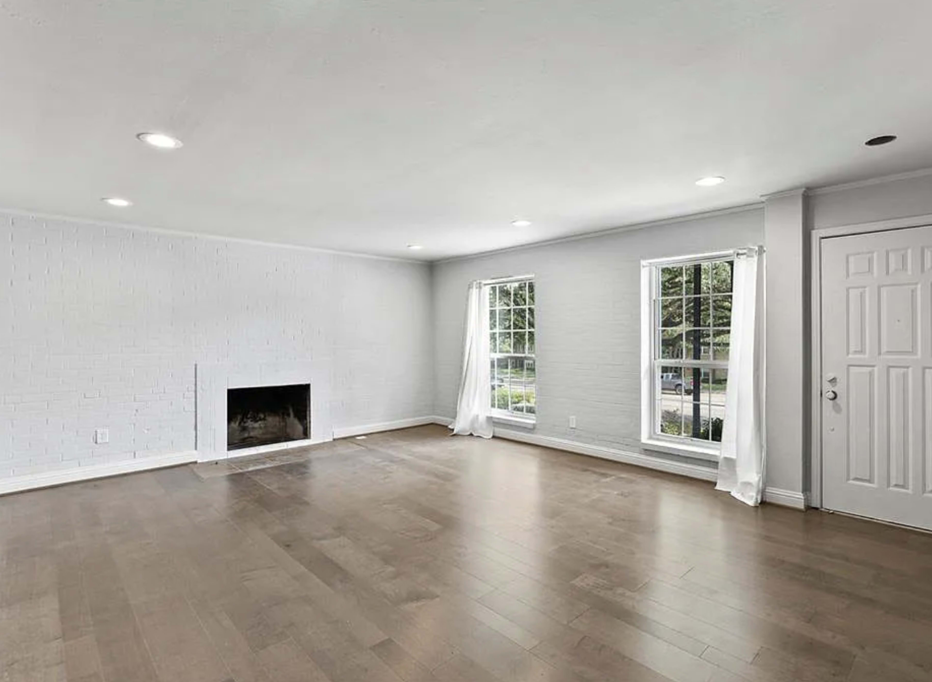 An empty living room with hardwood floors, large windows, and a brick fireplace.