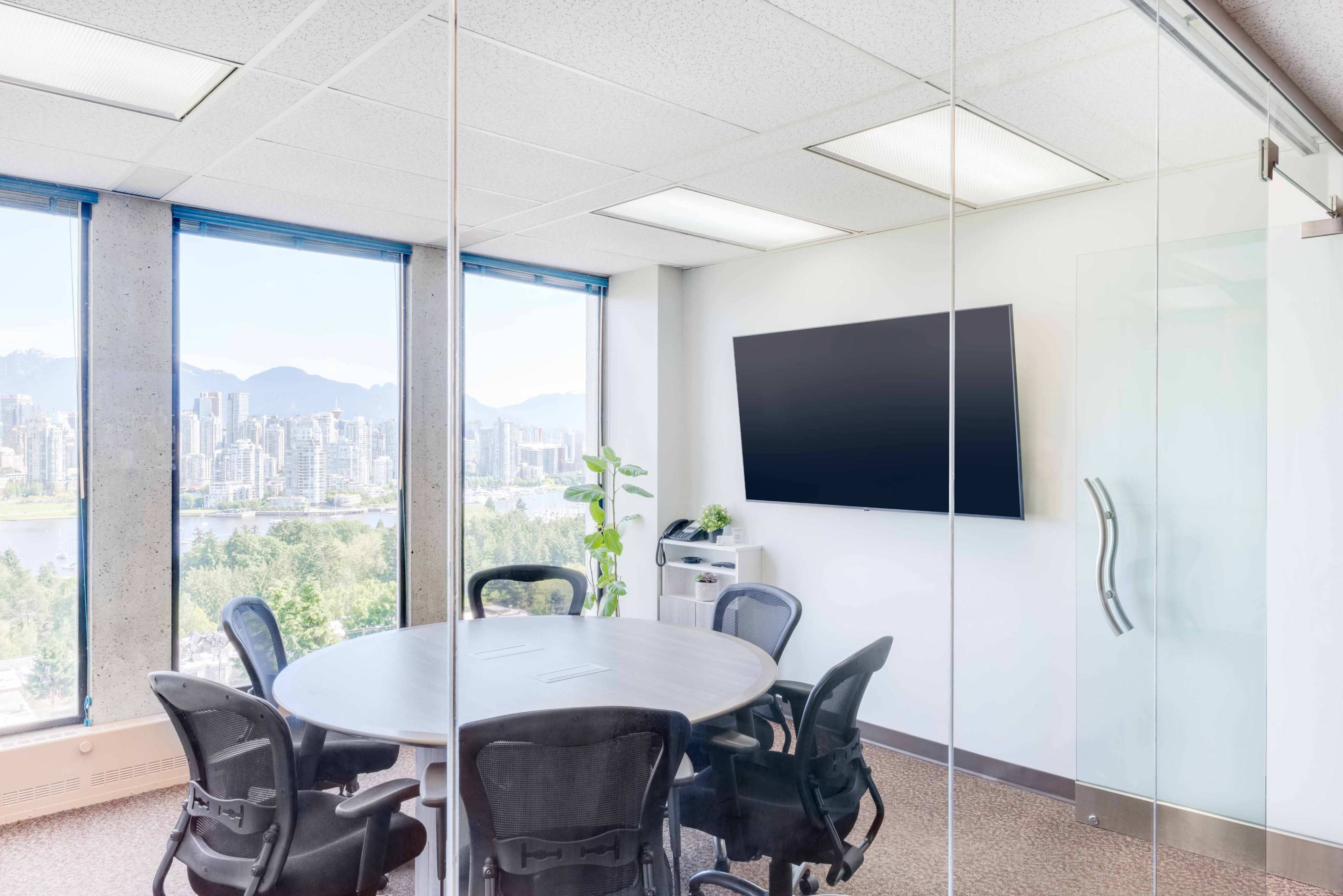 A modern meeting room features a circular table surrounded by chairs, with large windows offering a view of a city skyline and mountains in the background.