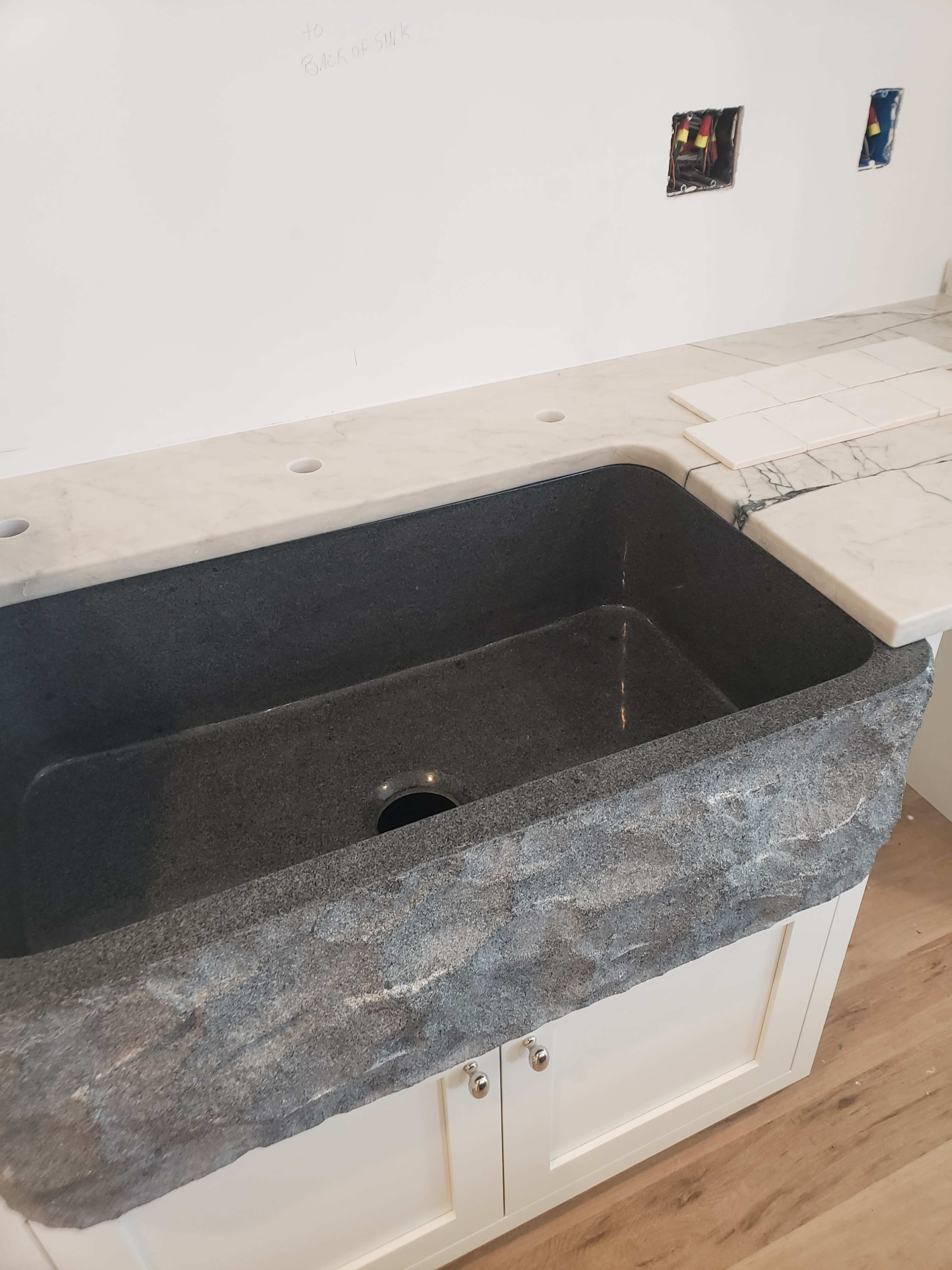 The image shows a dark granite farmhouse sink integrated into a light-colored countertop with a backsplash and unfinished wall electrical outlets visible.