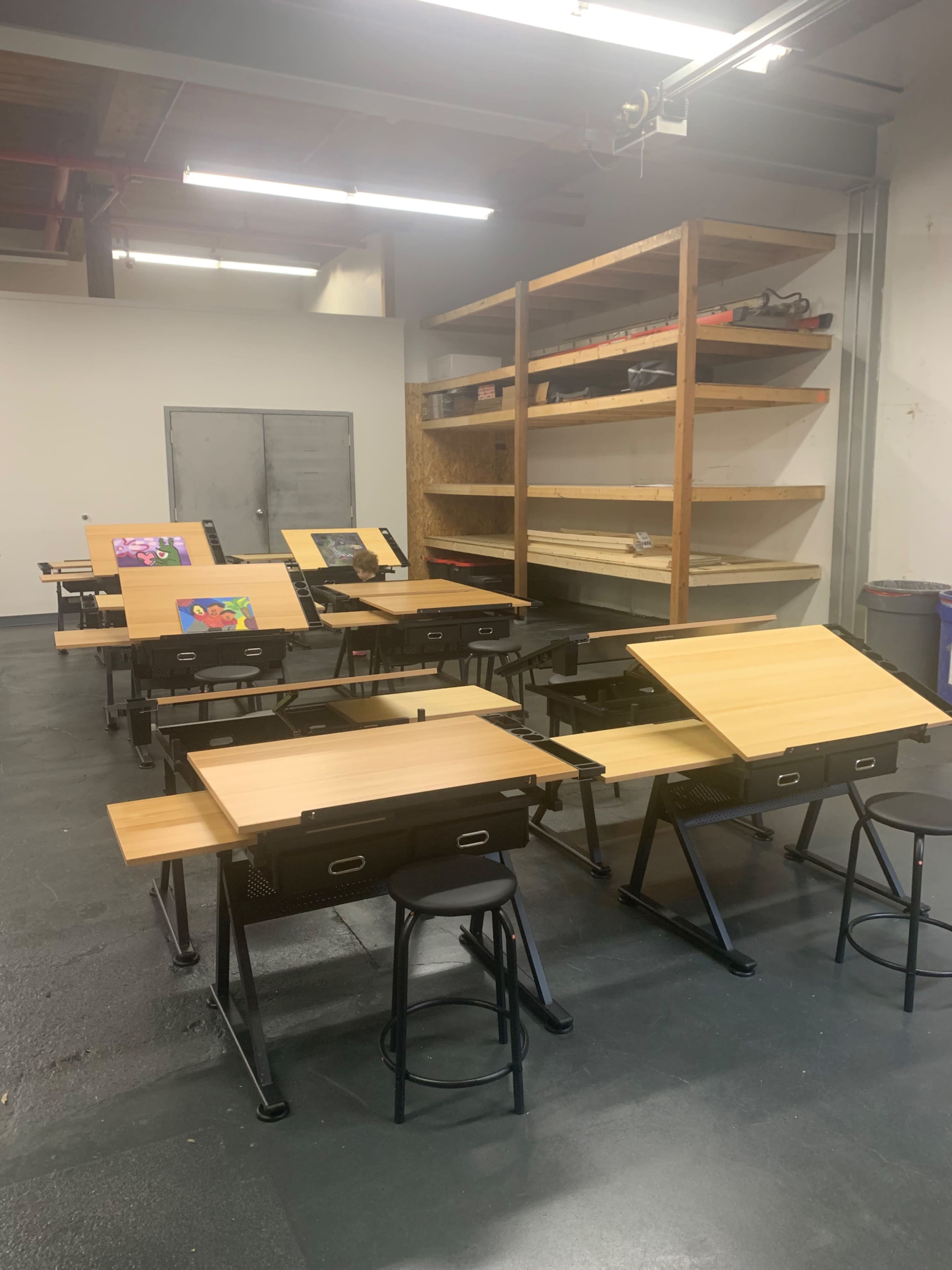 A room with multiple drafting tables and stools, arranged in rows, with shelves in the background.