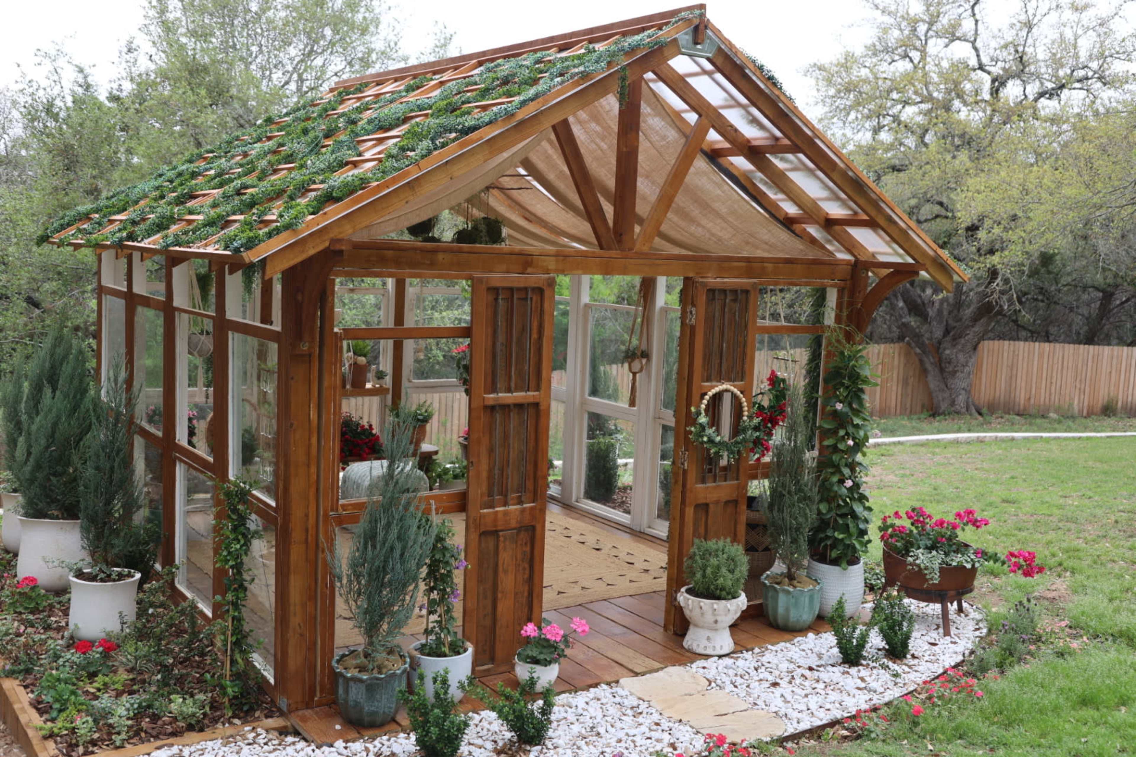 The image shows a wooden greenhouse surrounded by potted plants and flowers, featuring glass panels and a sloped roof.
