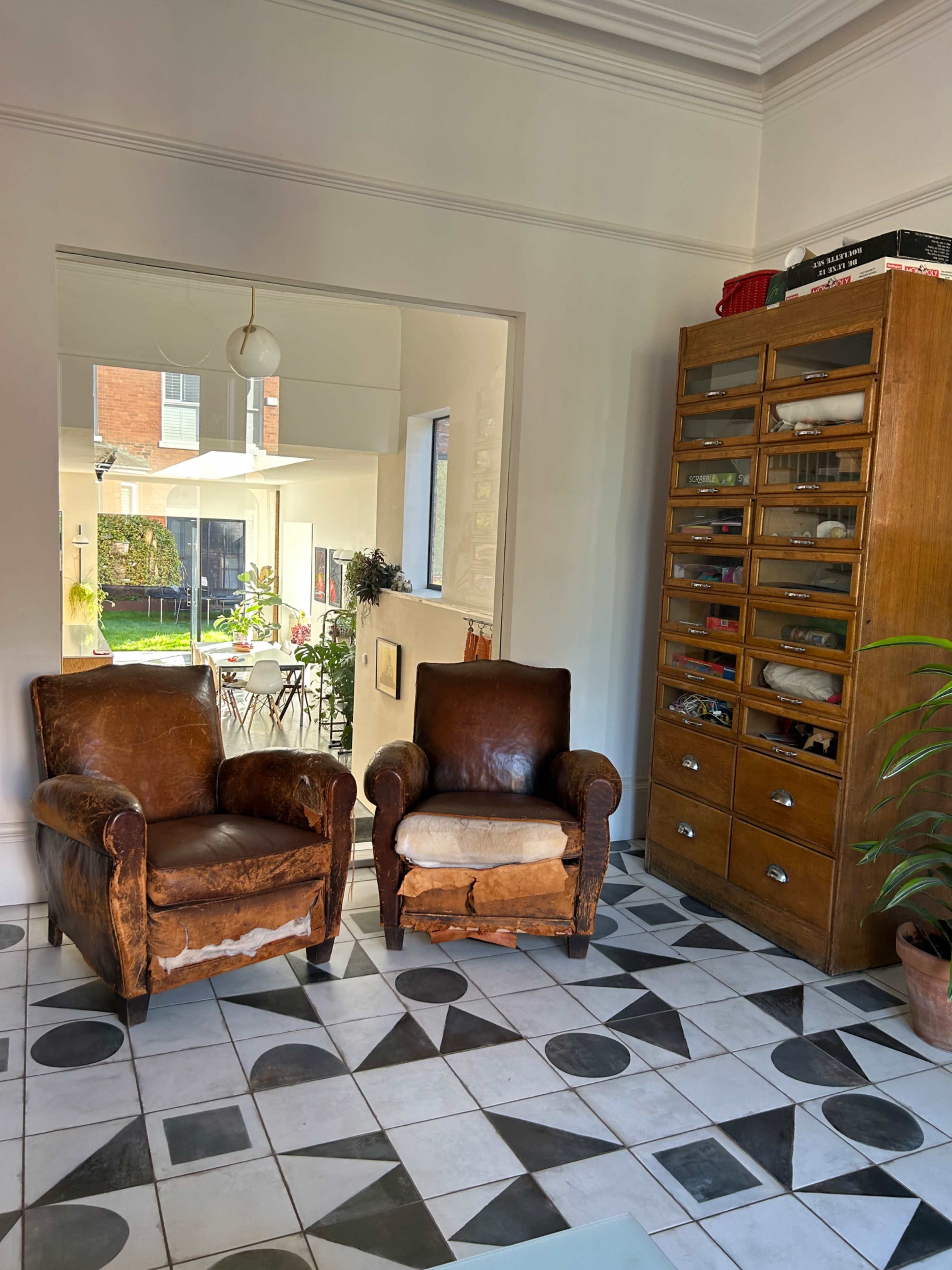 A room with two brown leather armchairs facing a wooden cabinet, and a light-filled area visible through an open doorway.
