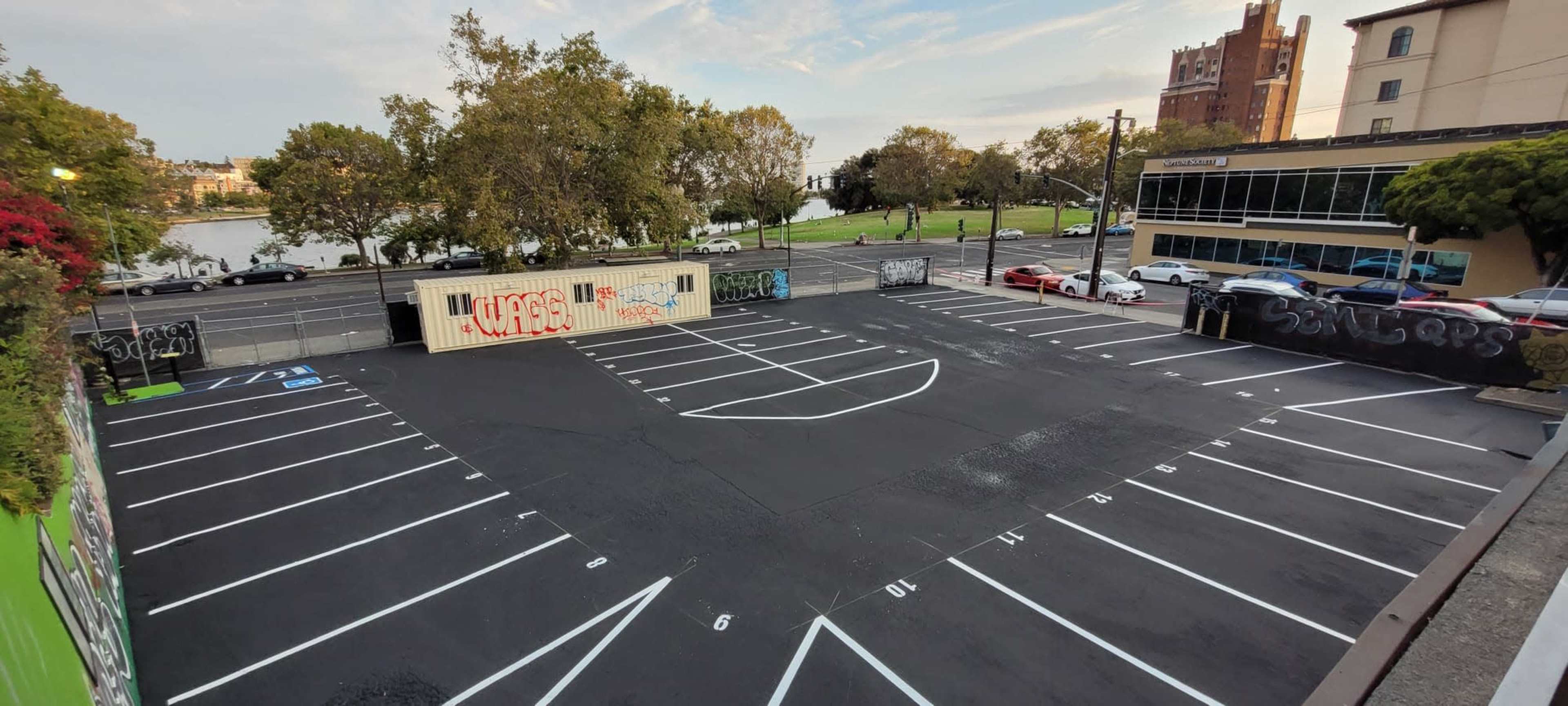 The image shows an empty parking lot with clearly marked parking spaces and a view of a nearby road and greenery in the background.