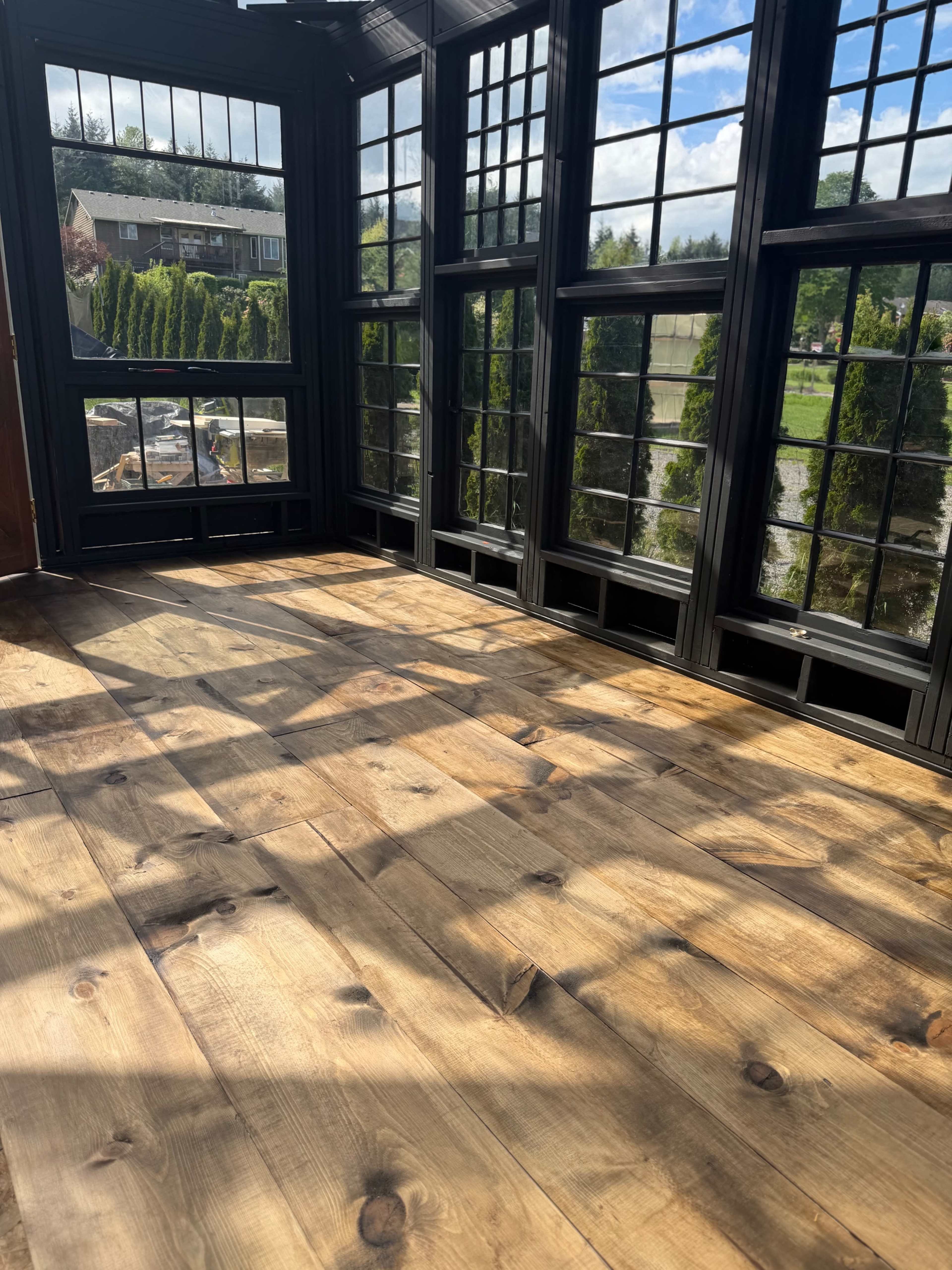 The image shows a sunlit room with wooden flooring and large black-framed windows offering a view of greenery outside.