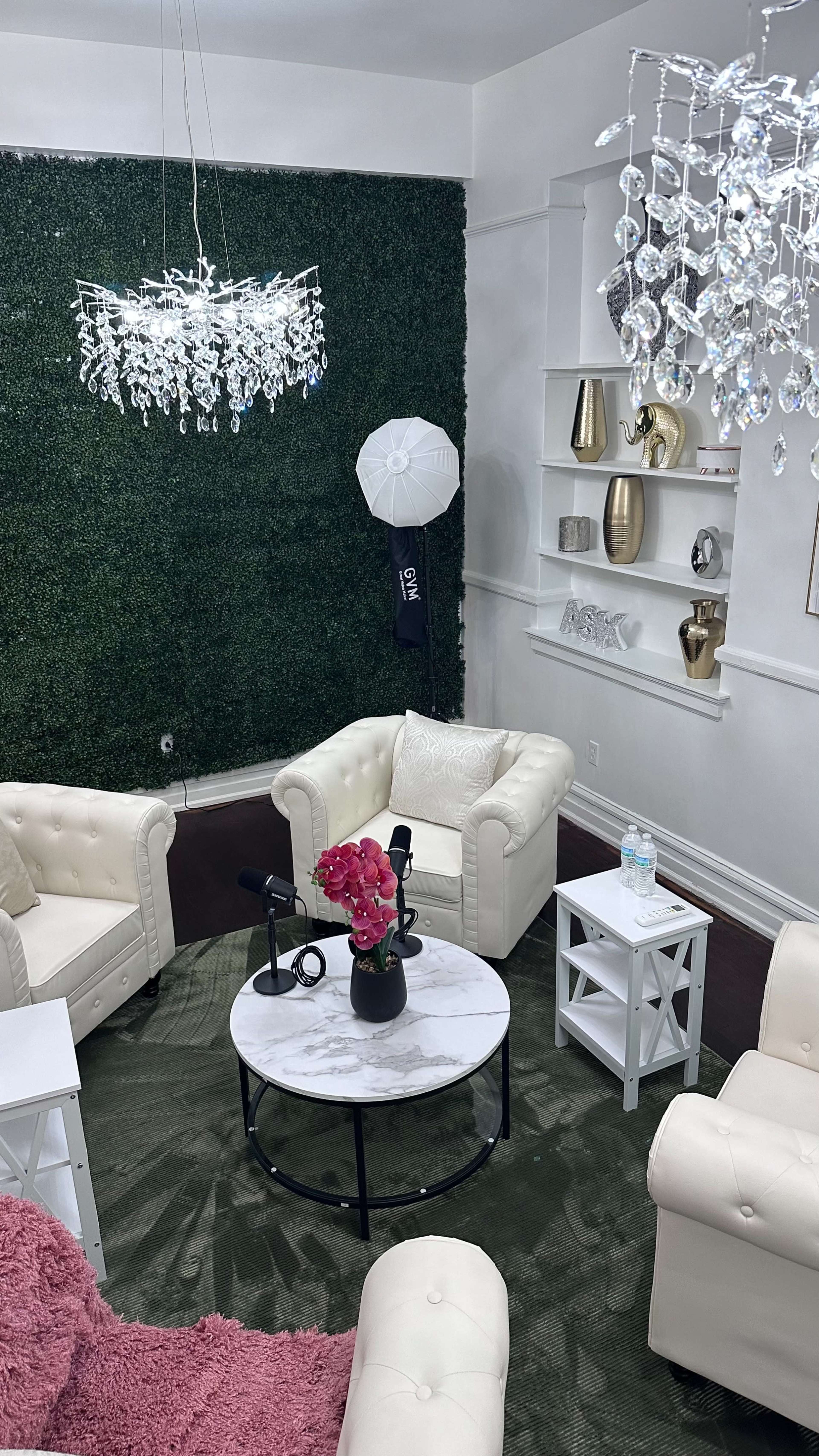 The image shows a modern living space with white chairs, a round marble table, decorative plants on the walls, and a chandelier.