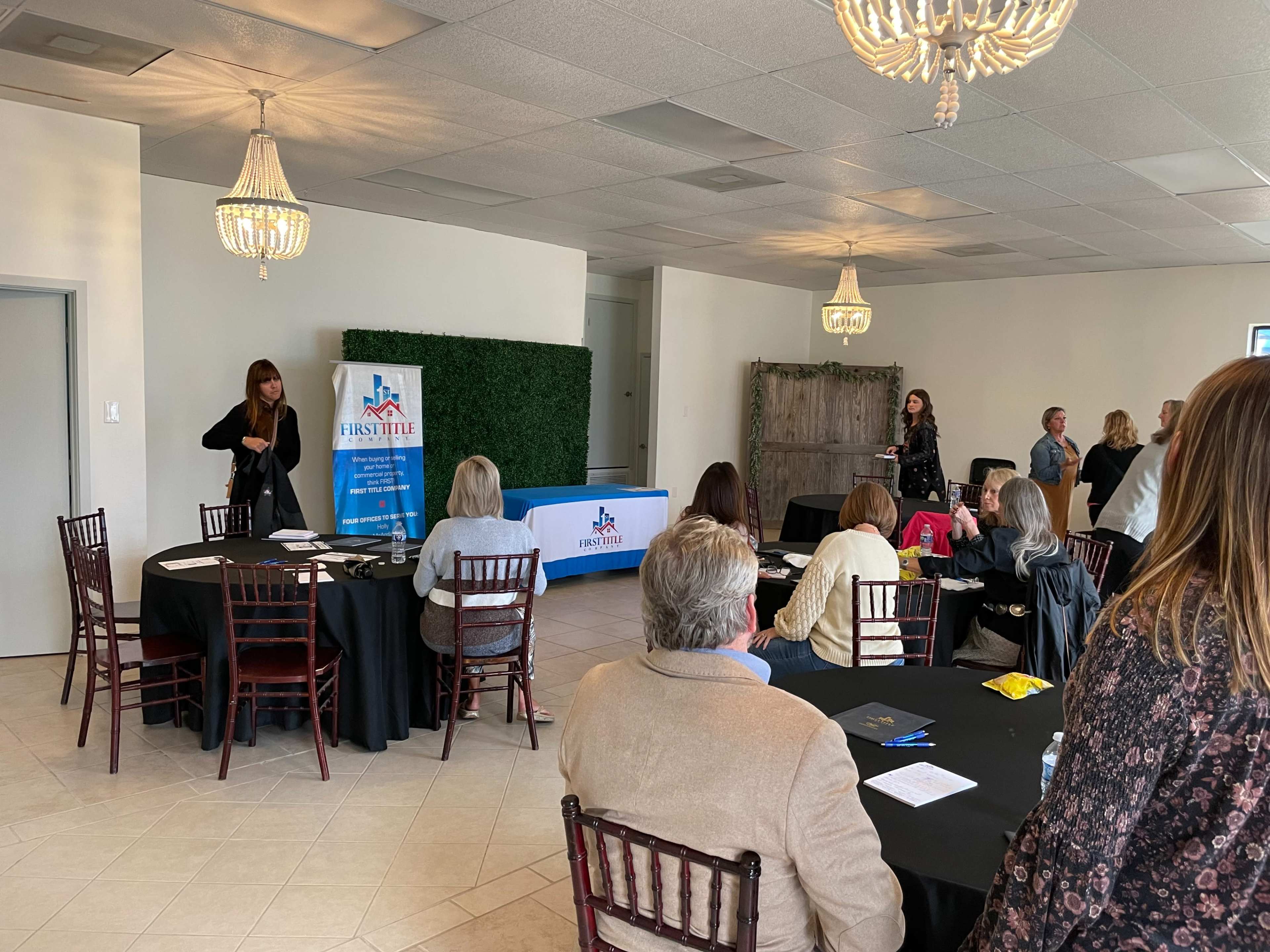 A group of people is seated at round tables in a well-lit room with a banner and a presentation area in the front.