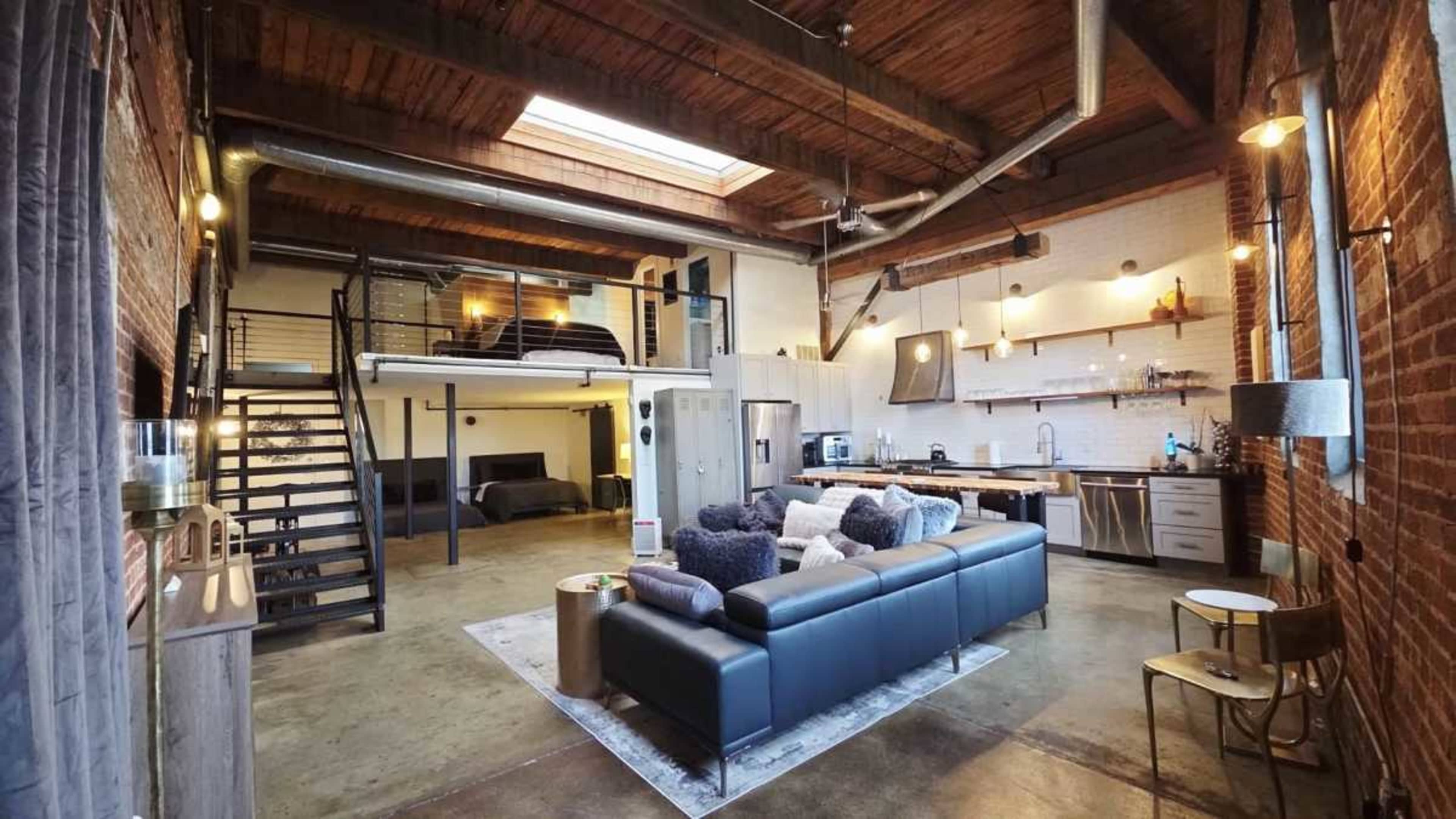 The image shows a spacious, modern loft with exposed brick walls, a large sectional sofa, a kitchen area, and a staircase leading to a sleeping loft.