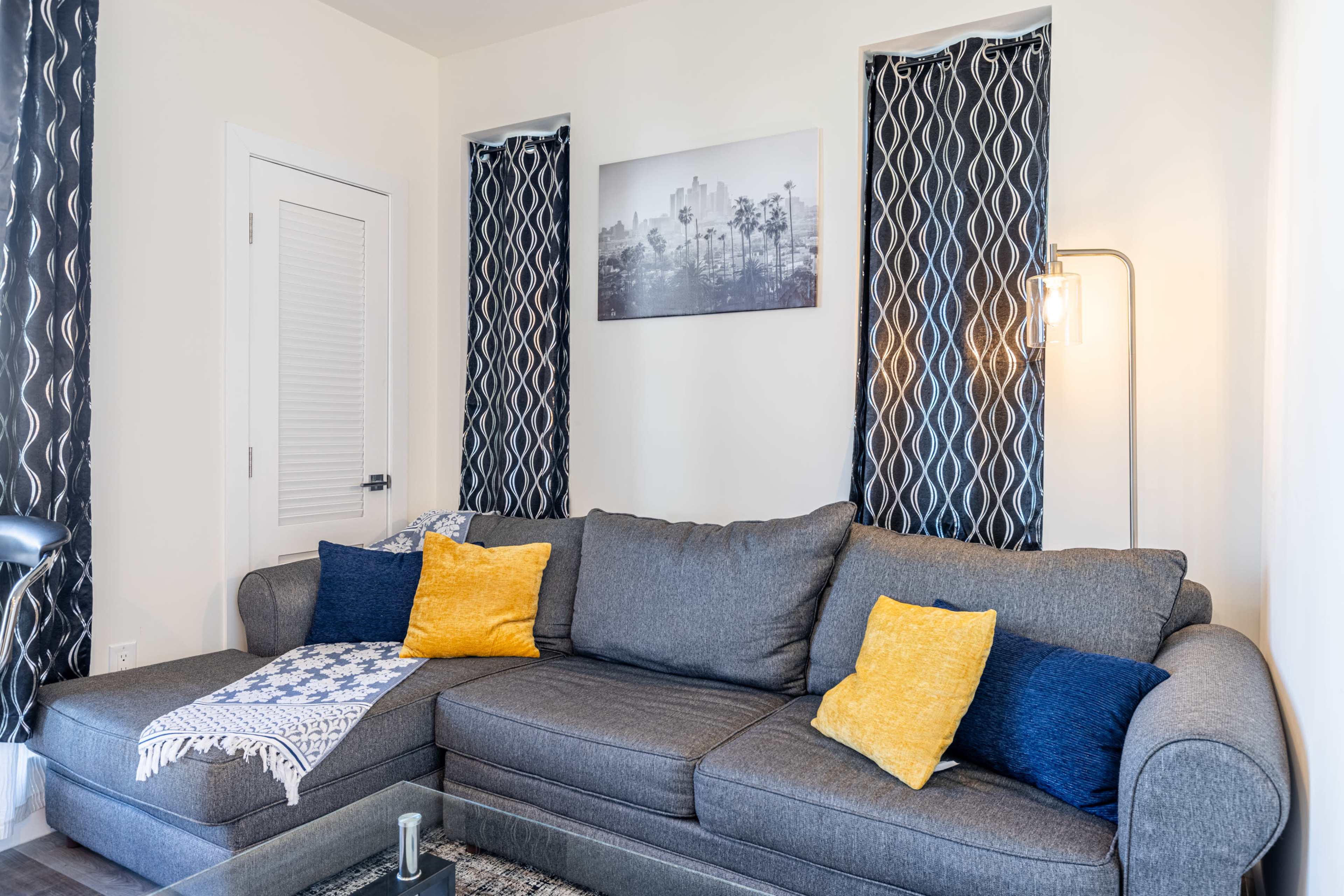The image shows a modern living room with a gray sofa adorned with yellow and blue throw pillows, surrounded by patterned curtains and a wall featuring a black-and-white cityscape photograph.