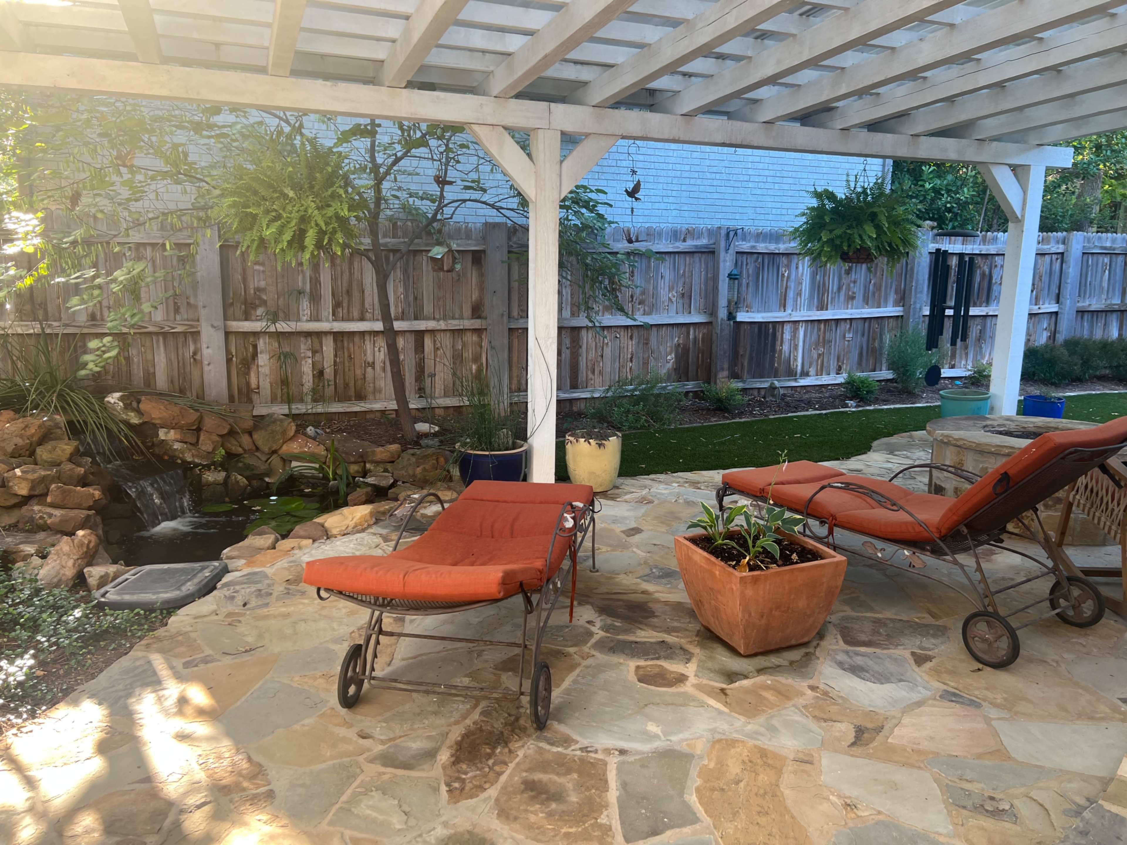 A backyard patio with two orange lounge chairs, a stone floor, and a small water feature surrounded by greenery and potted plants.