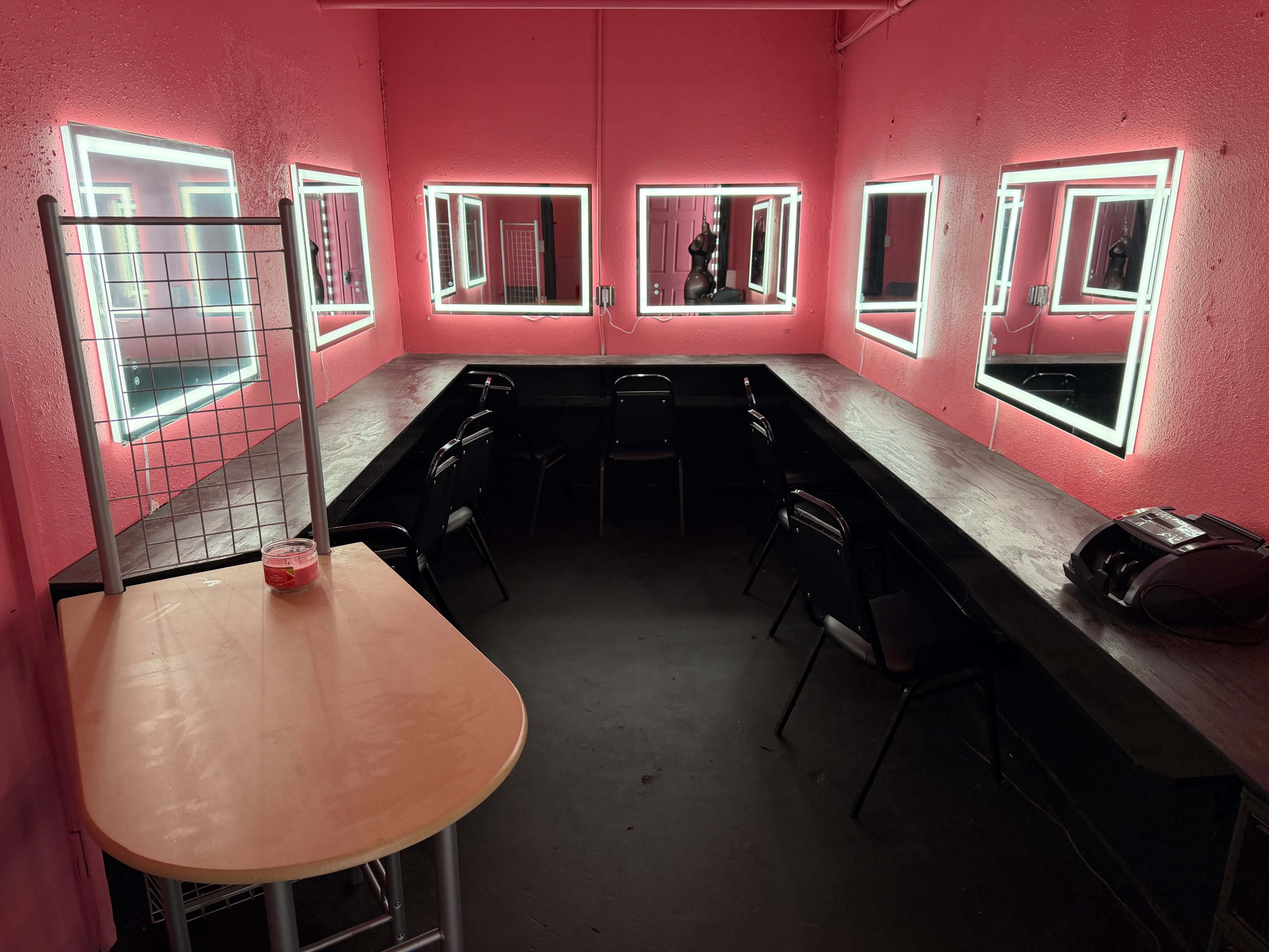 The room features bright pink walls, several illuminated mirrors, and a long dark table surrounded by black chairs.