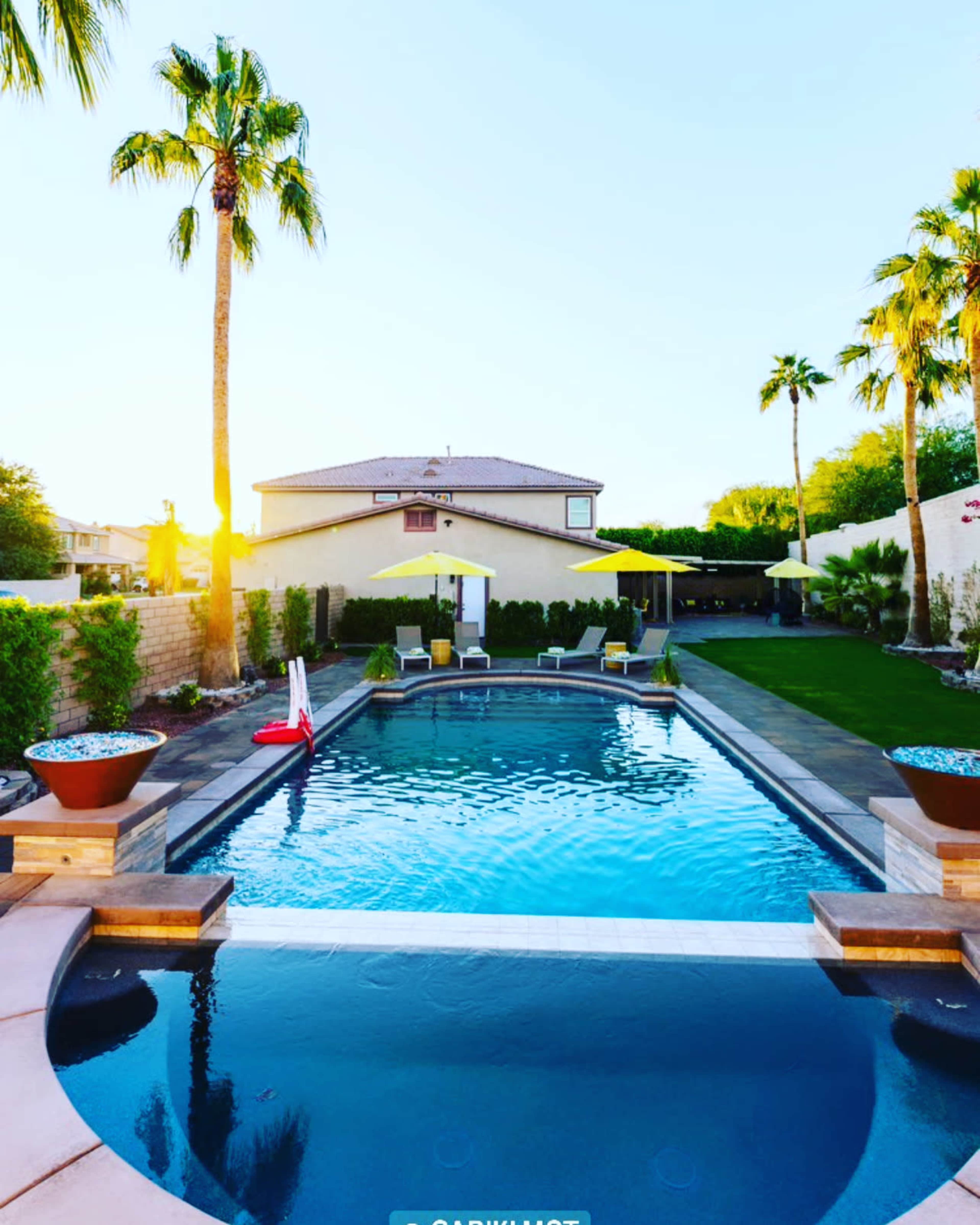 The image shows a clear swimming pool surrounded by palm trees and lounge chairs, with a house in the background and a bright sunset.