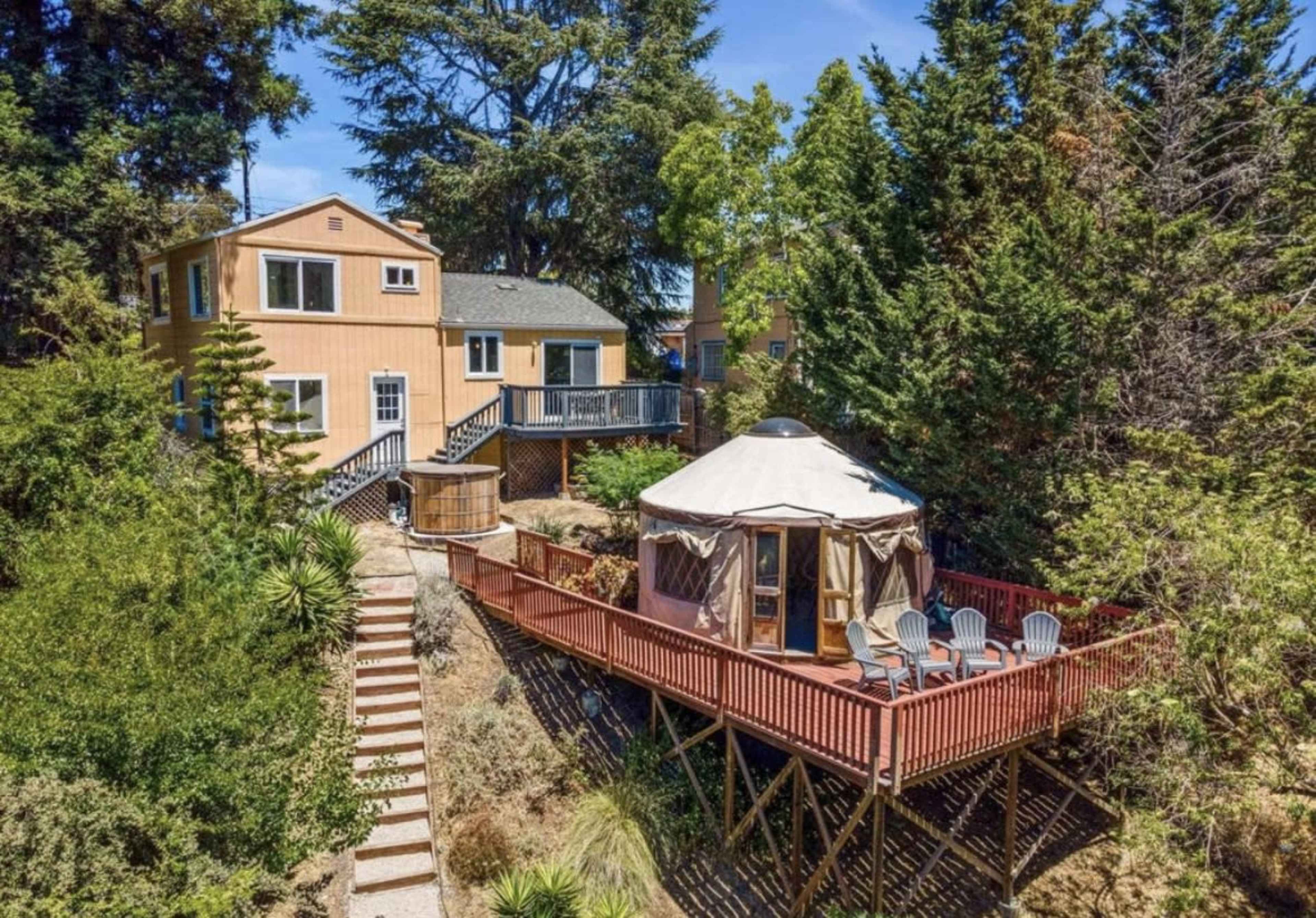 The image shows a two-story house and a yurt on a wooden deck surrounded by trees and vegetation.
