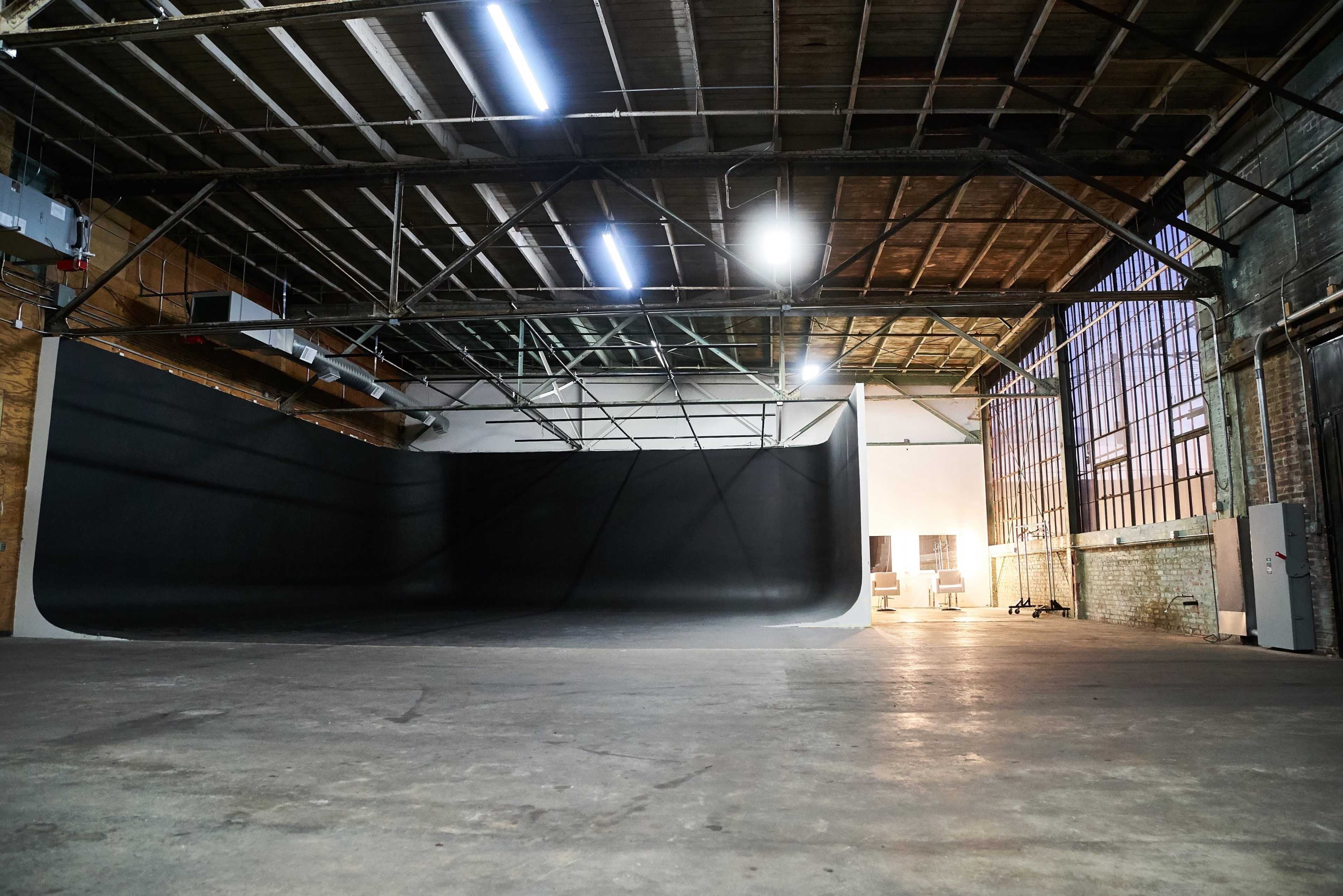 The image shows a large, empty industrial studio space with a black backdrop and exposed beams.