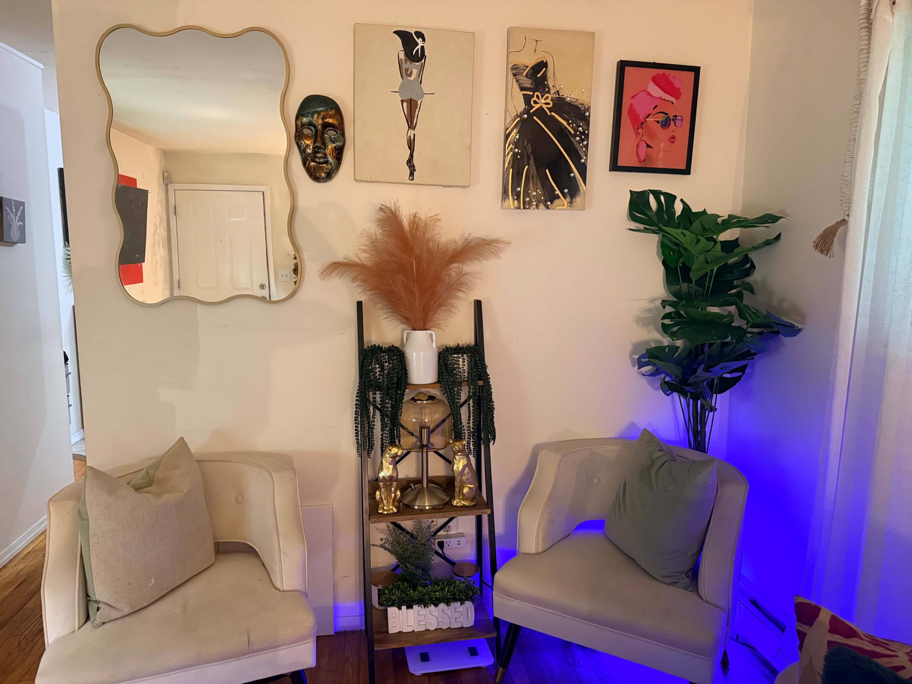 The image shows a cozy corner of a living room featuring two cream-colored chairs, a decorative shelf with plants and gold accents, and a wall adorned with art pieces and a mirror.