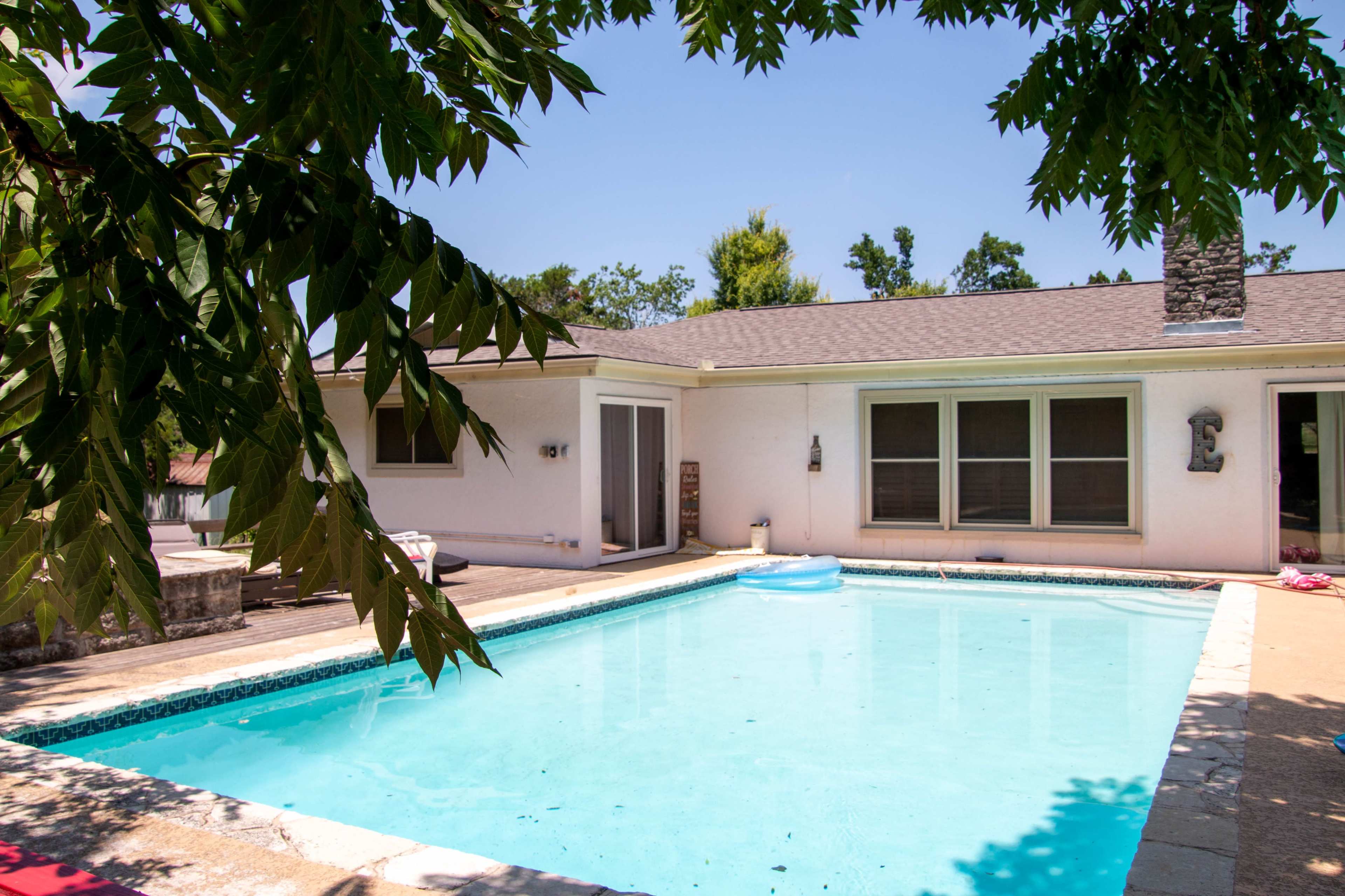 A clear swimming pool surrounded by a patio and a house with large windows on a sunny day.