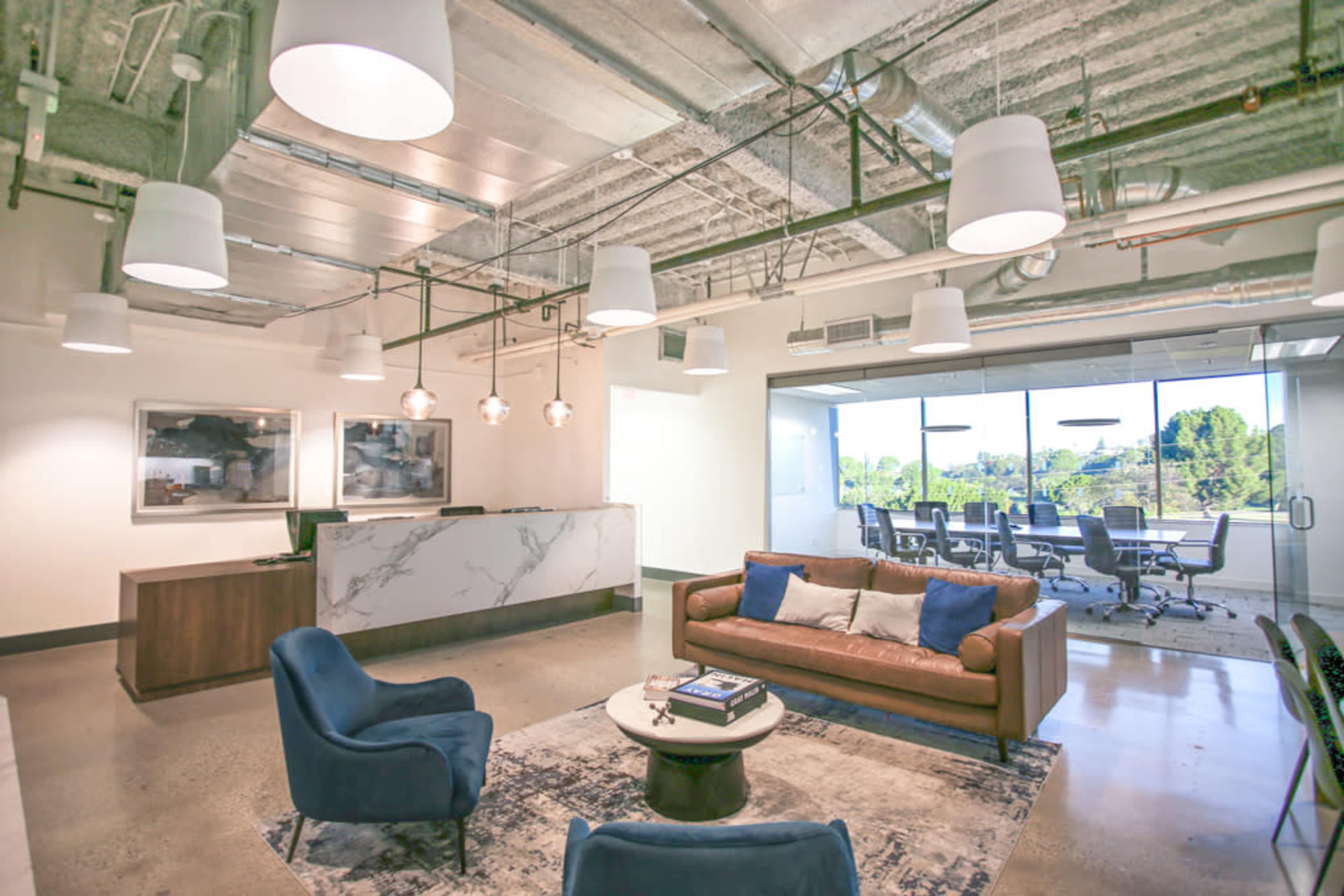 A modern office lobby with a reception desk, a seating area featuring a brown couch and blue chairs, and a glass-walled meeting room in the background.