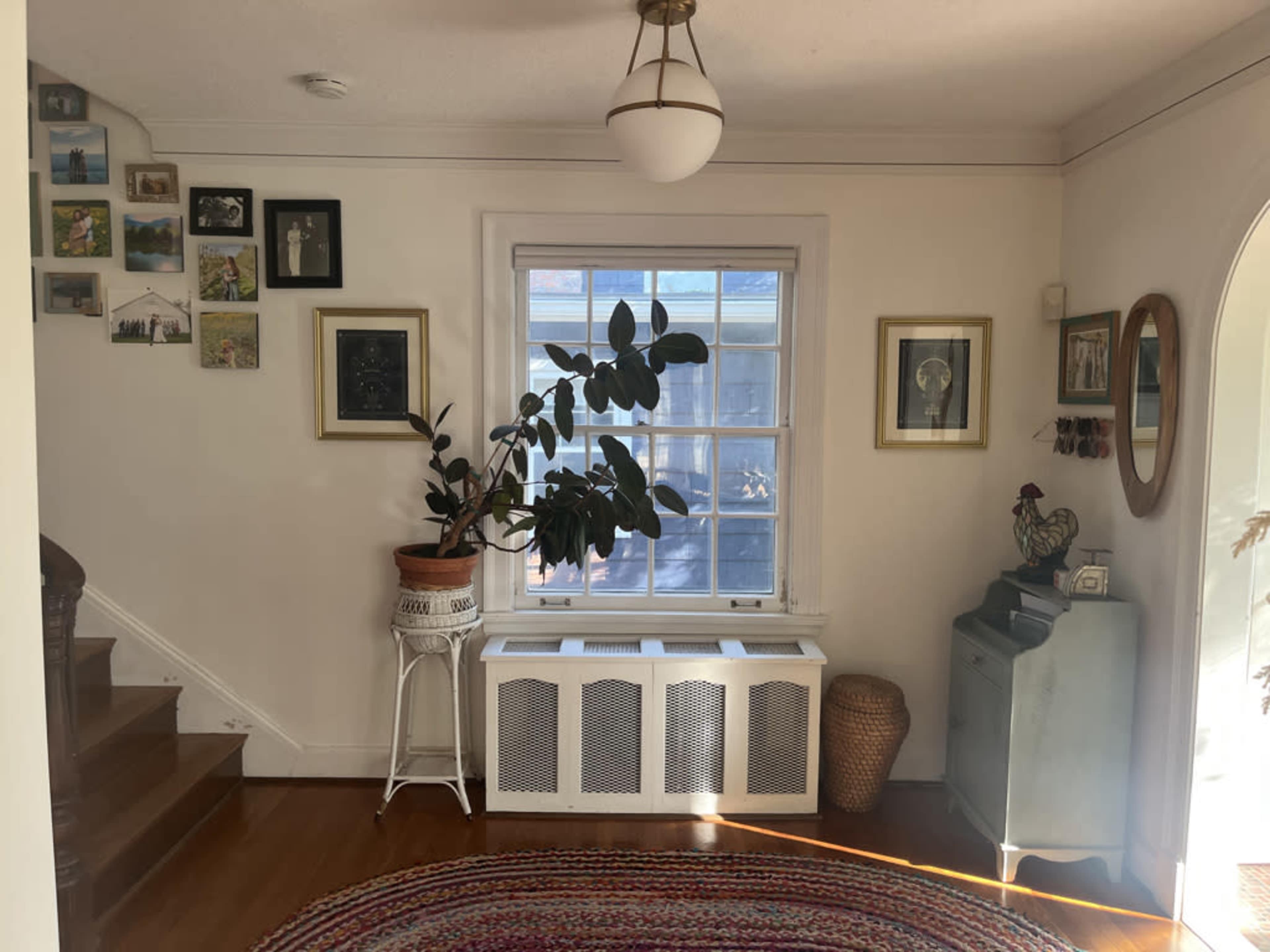 The image shows a residential hallway with a staircase on the left, a window illuminated by natural light, and various framed pictures on the walls.