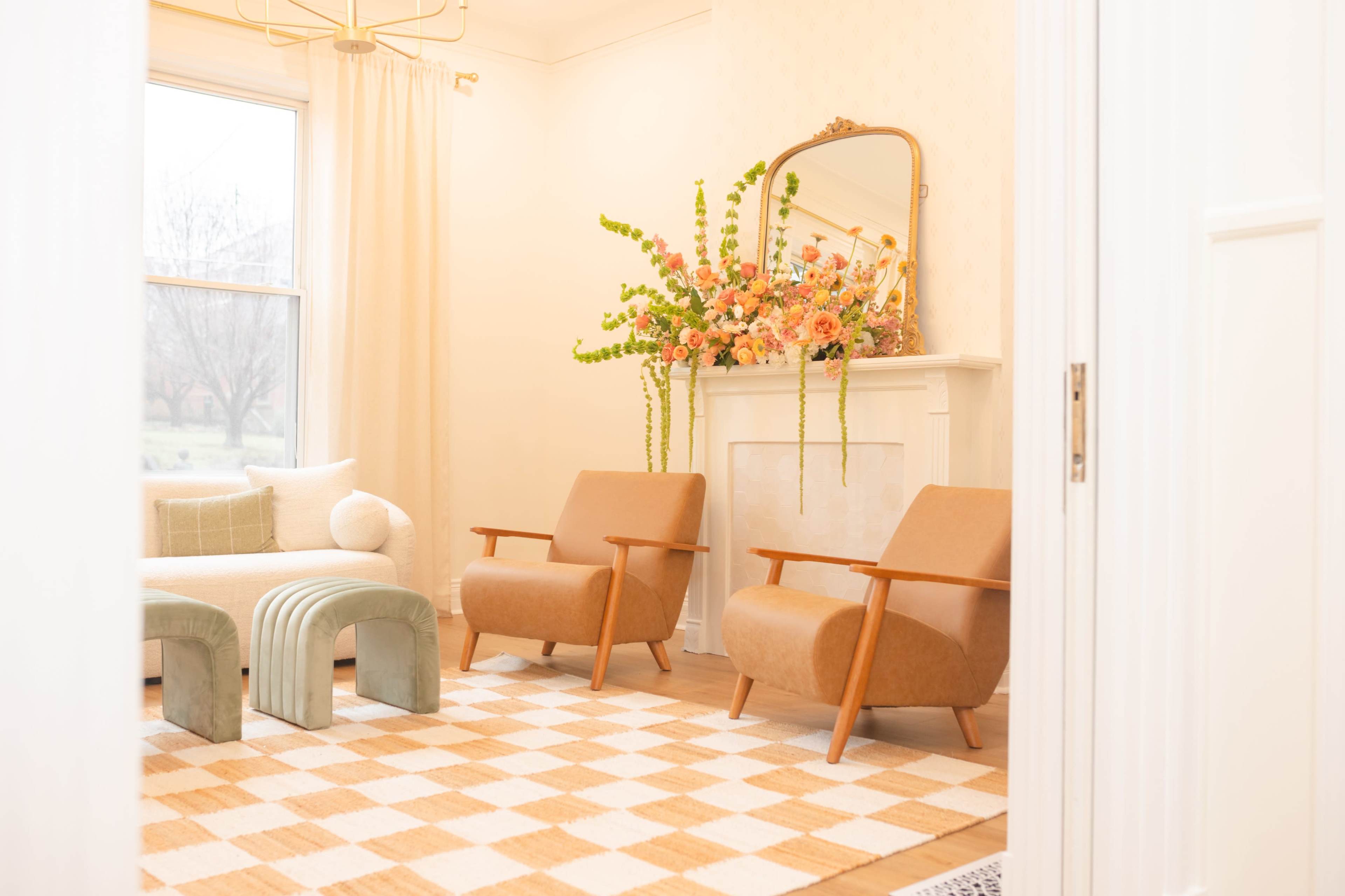 The room features two brown armchairs, a green curved bench, a checkered rug, and a mantel decorated with a floral arrangement.