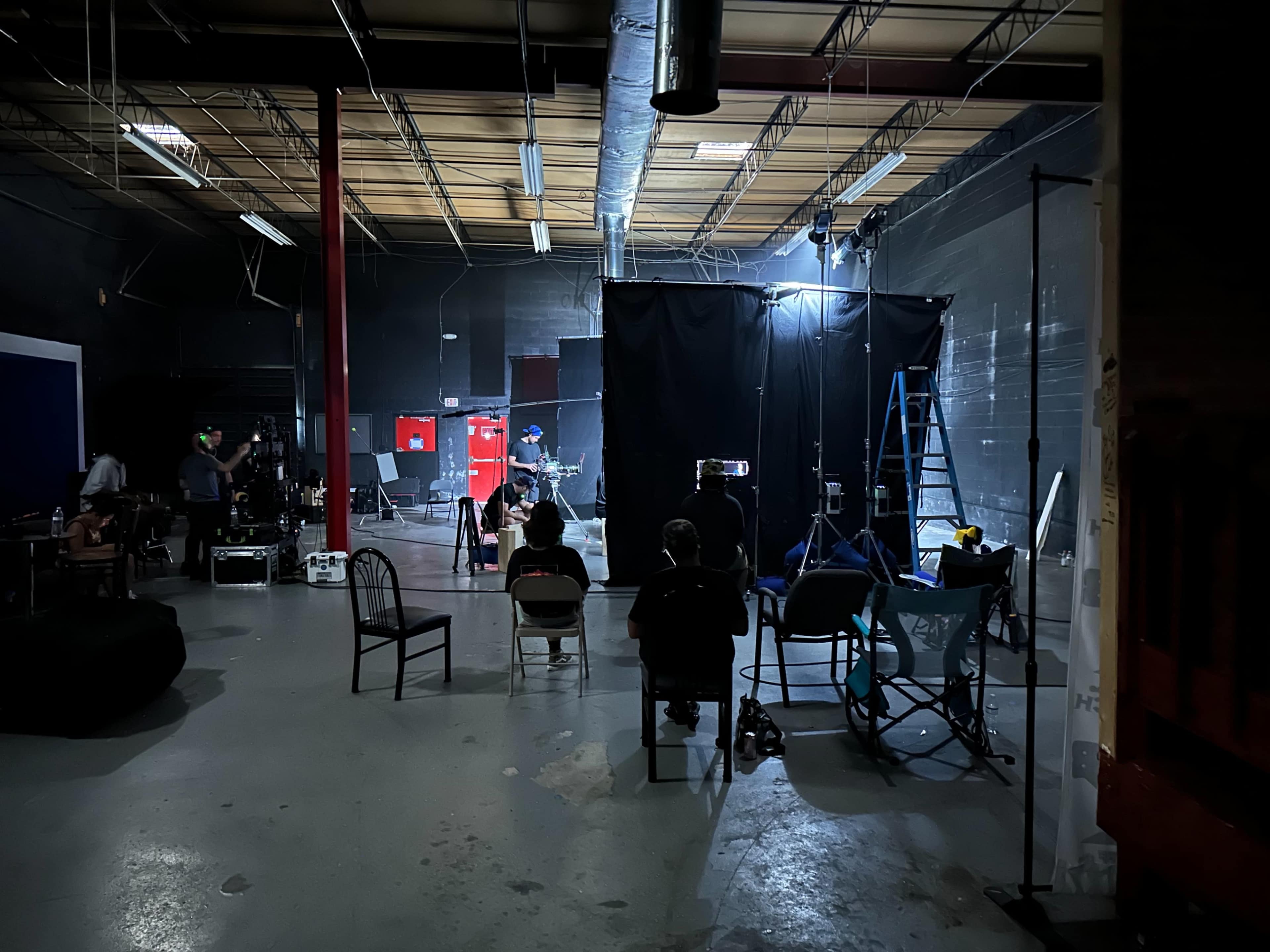 A group of people sits on chairs in a dimly lit studio, facing a setup with lights and a black backdrop.