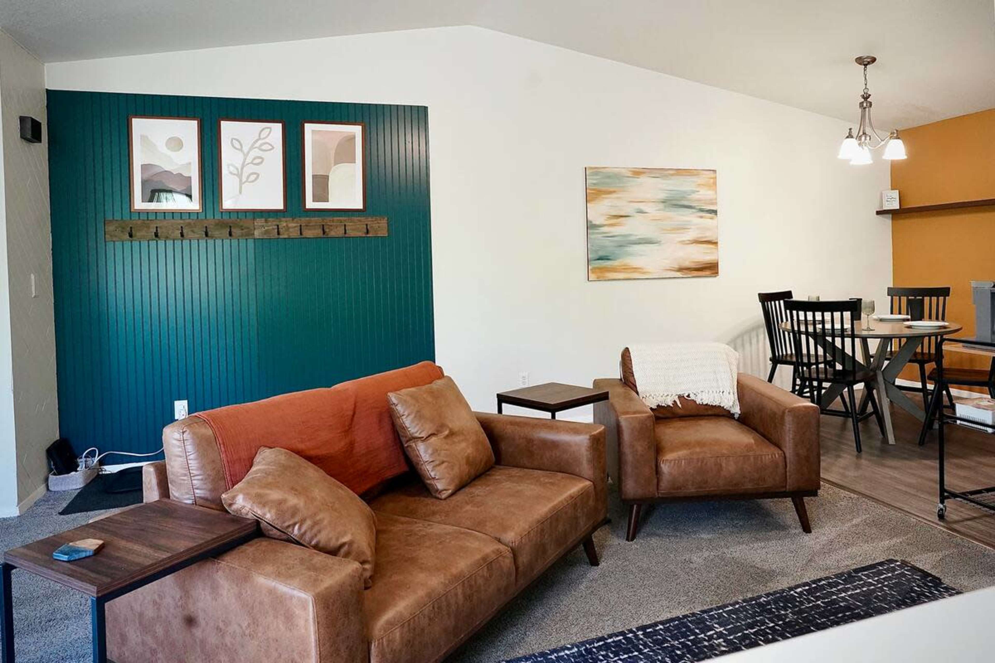 The image shows a modern living space featuring a brown leather sofa, a green accent wall with framed artwork, and a dining area with a table and chairs.