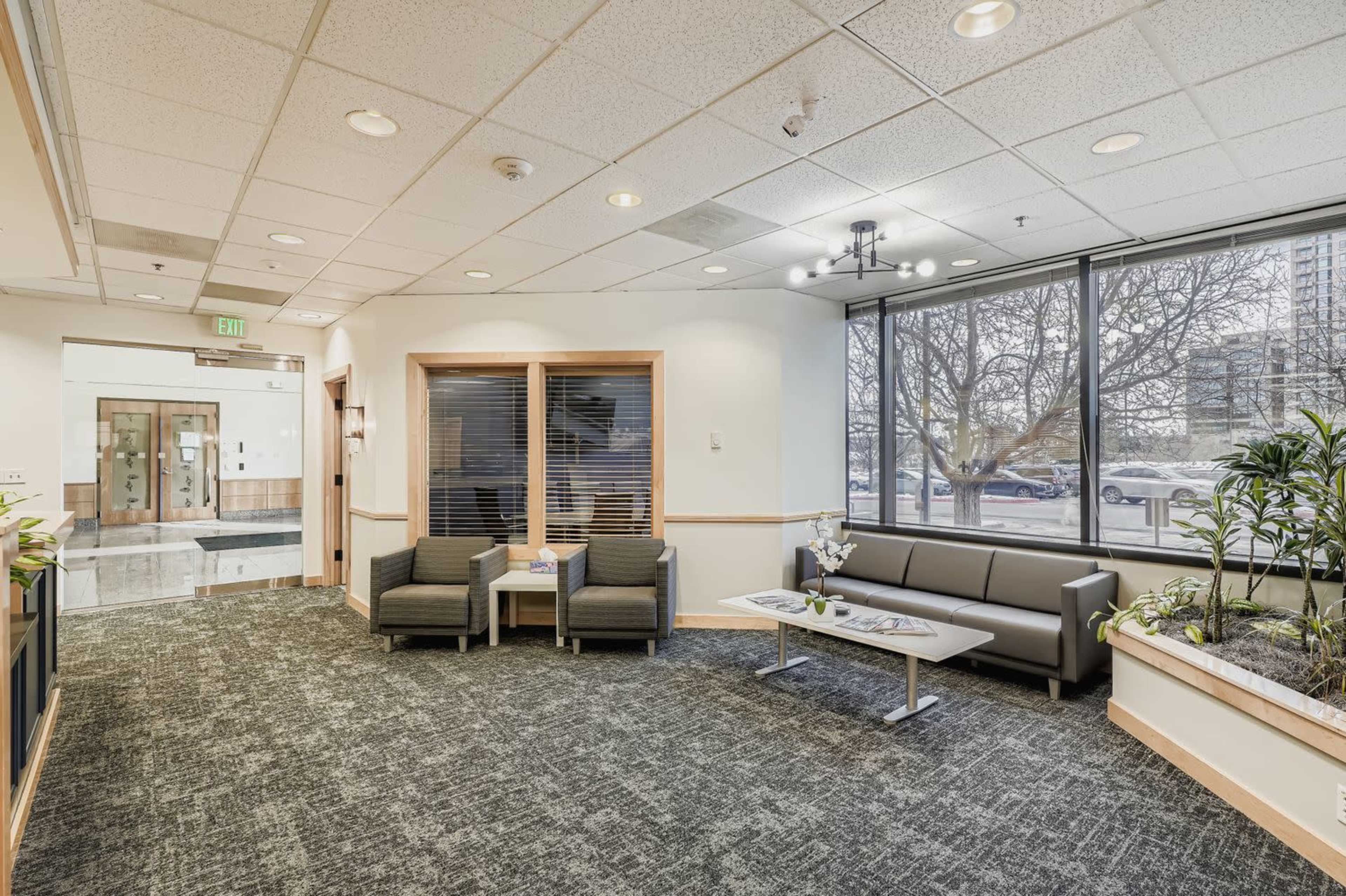 The image shows a modern waiting area featuring gray seating, large windows with natural light, and plants along the walls.