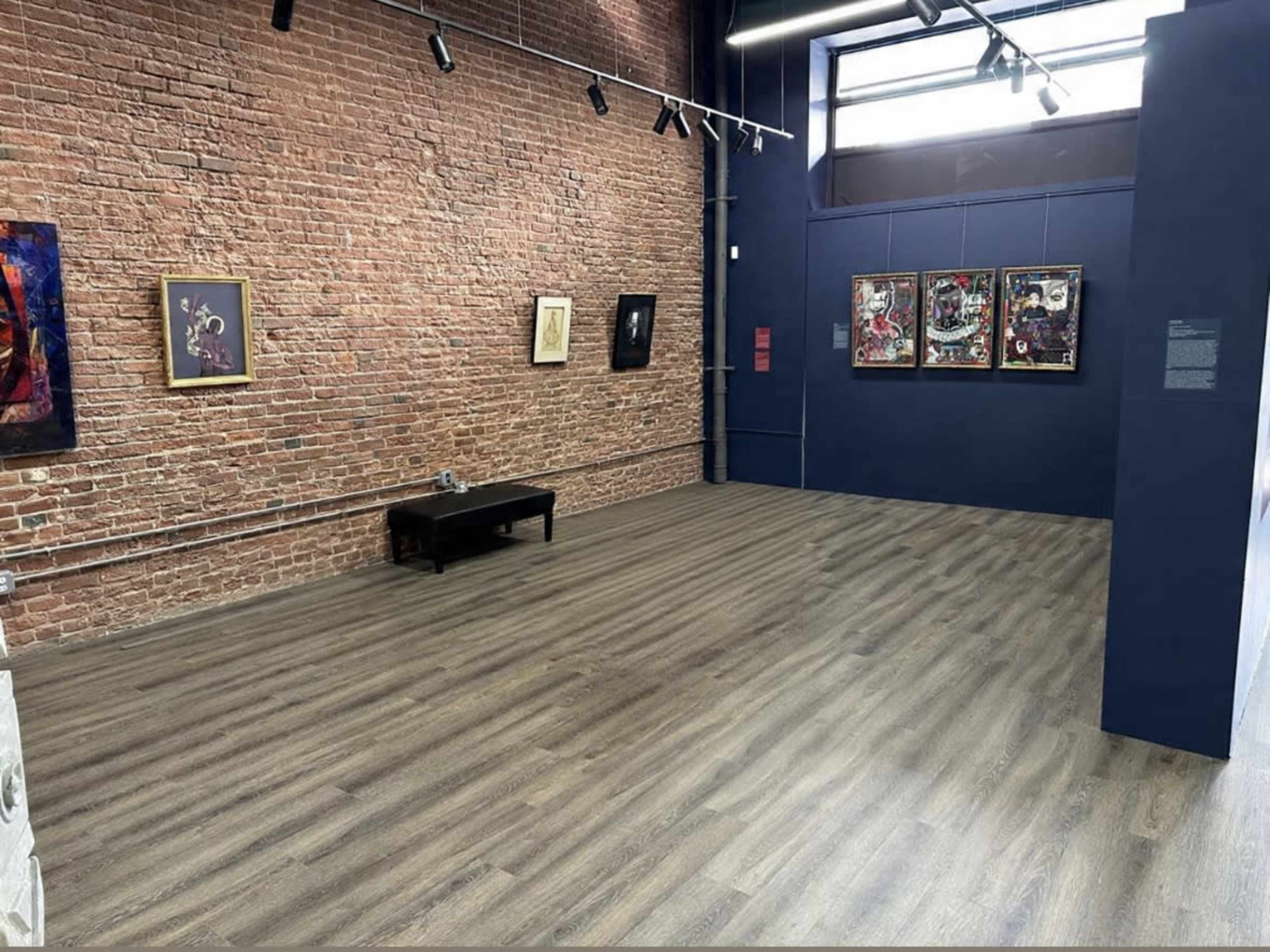 The image shows a spacious art gallery featuring exposed brick walls and wooden flooring, with several framed artworks displayed on one wall.