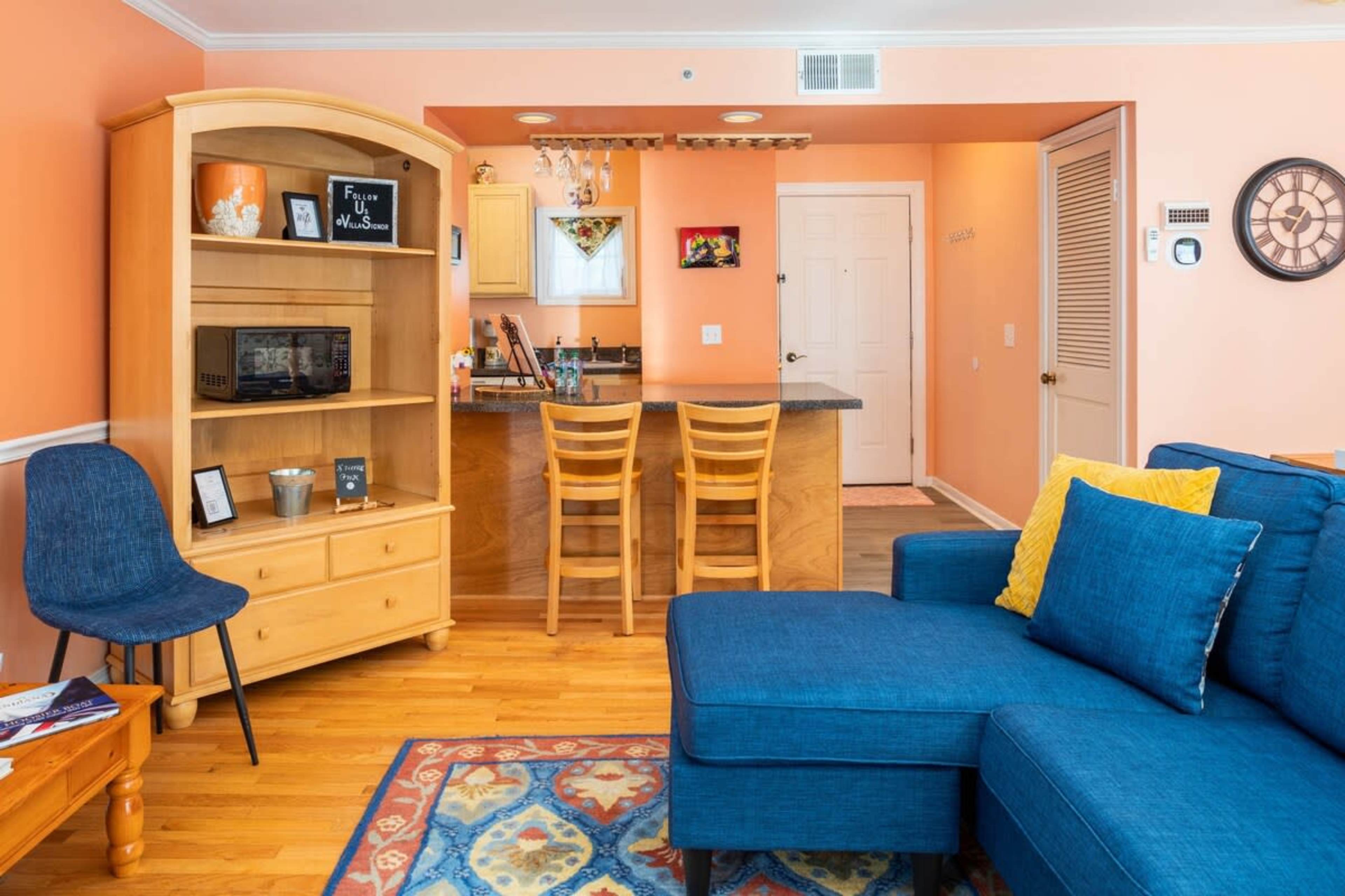 The image shows a cozy living space with an orange wall, a blue sectional sofa, a wooden shelf unit, and a kitchen area in the background.