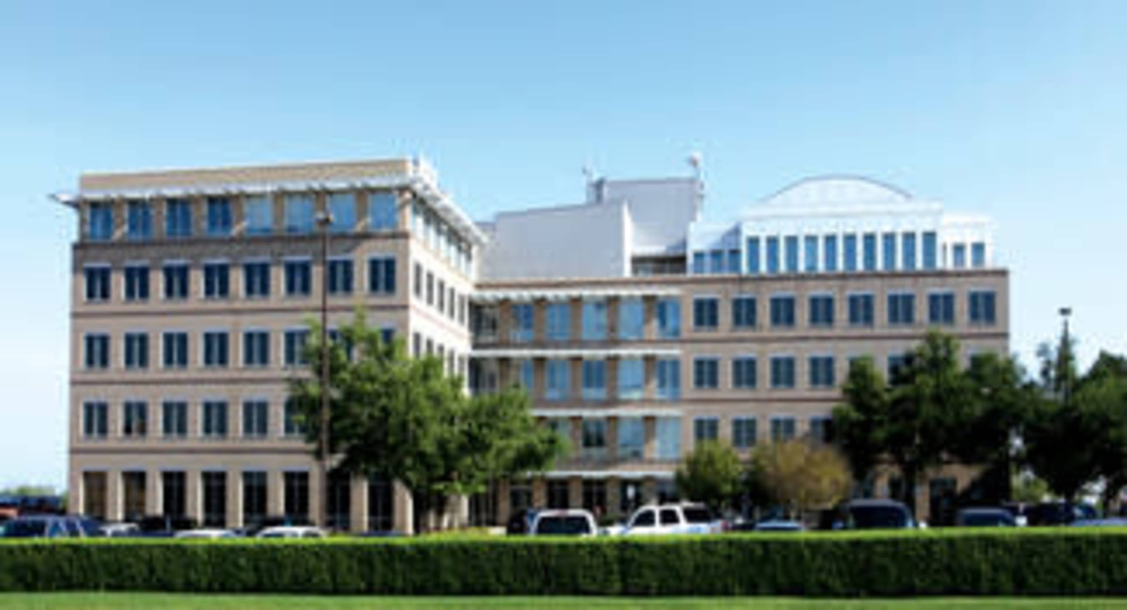 The image shows a multi-story office building with large windows and a flat roof, surrounded by a landscaped area and parked cars.