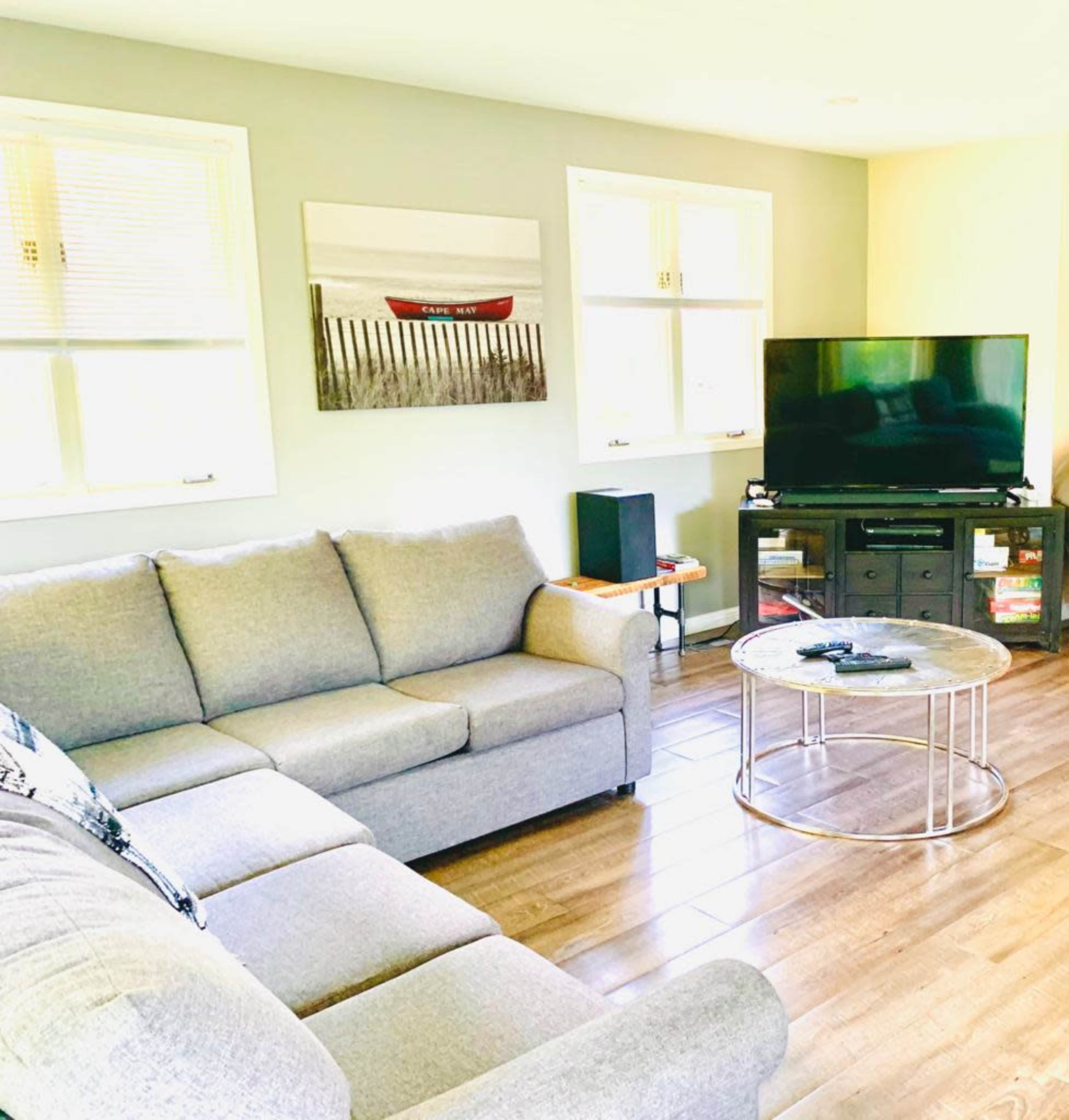 A living room features a gray sectional sofa, a glass coffee table, and a TV on a stand, with light streaming in from windows.