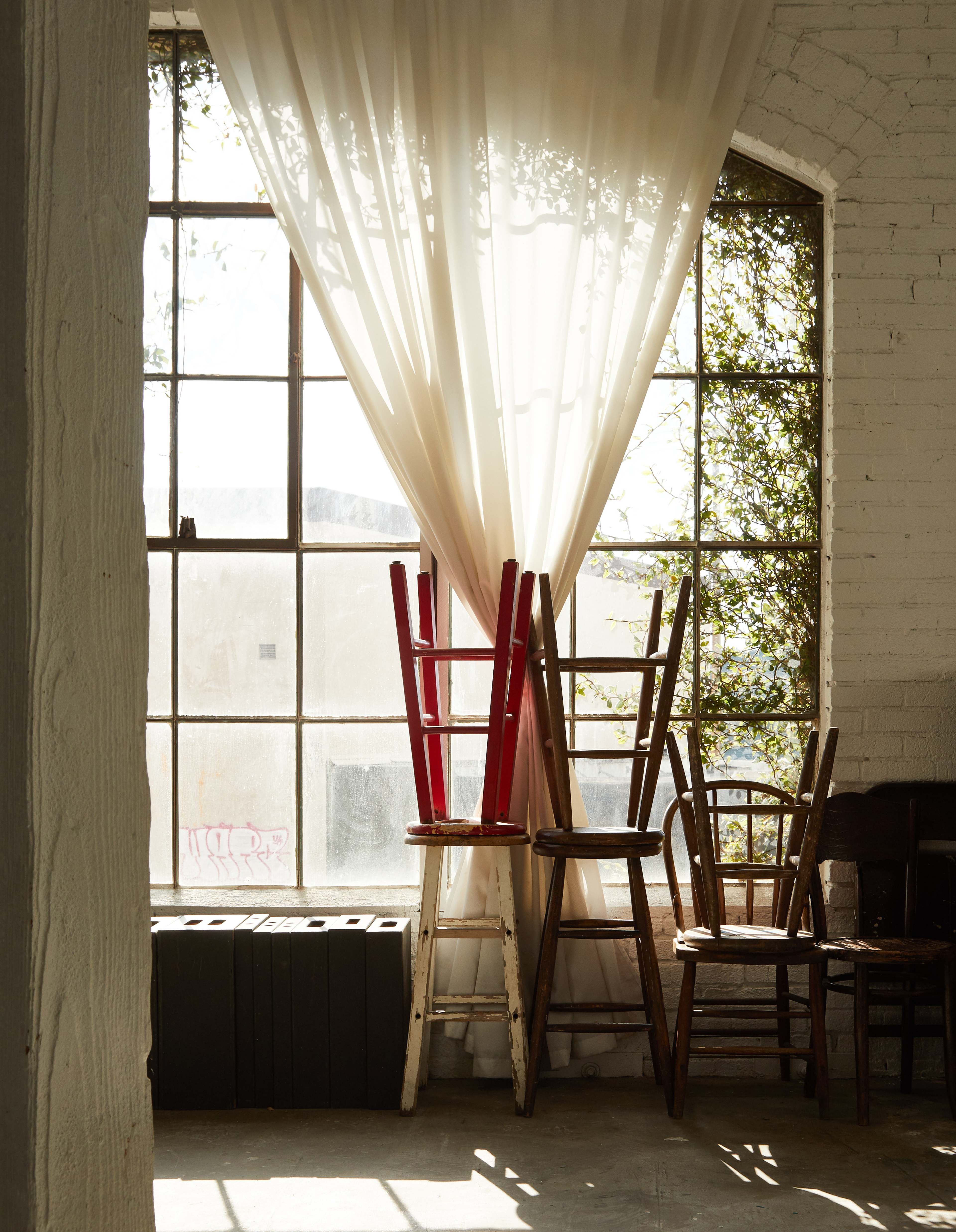 Two wooden chairs are stacked leaning against a large window with sheer curtains, alongside another chair placed on the floor in a room filled with natural light.