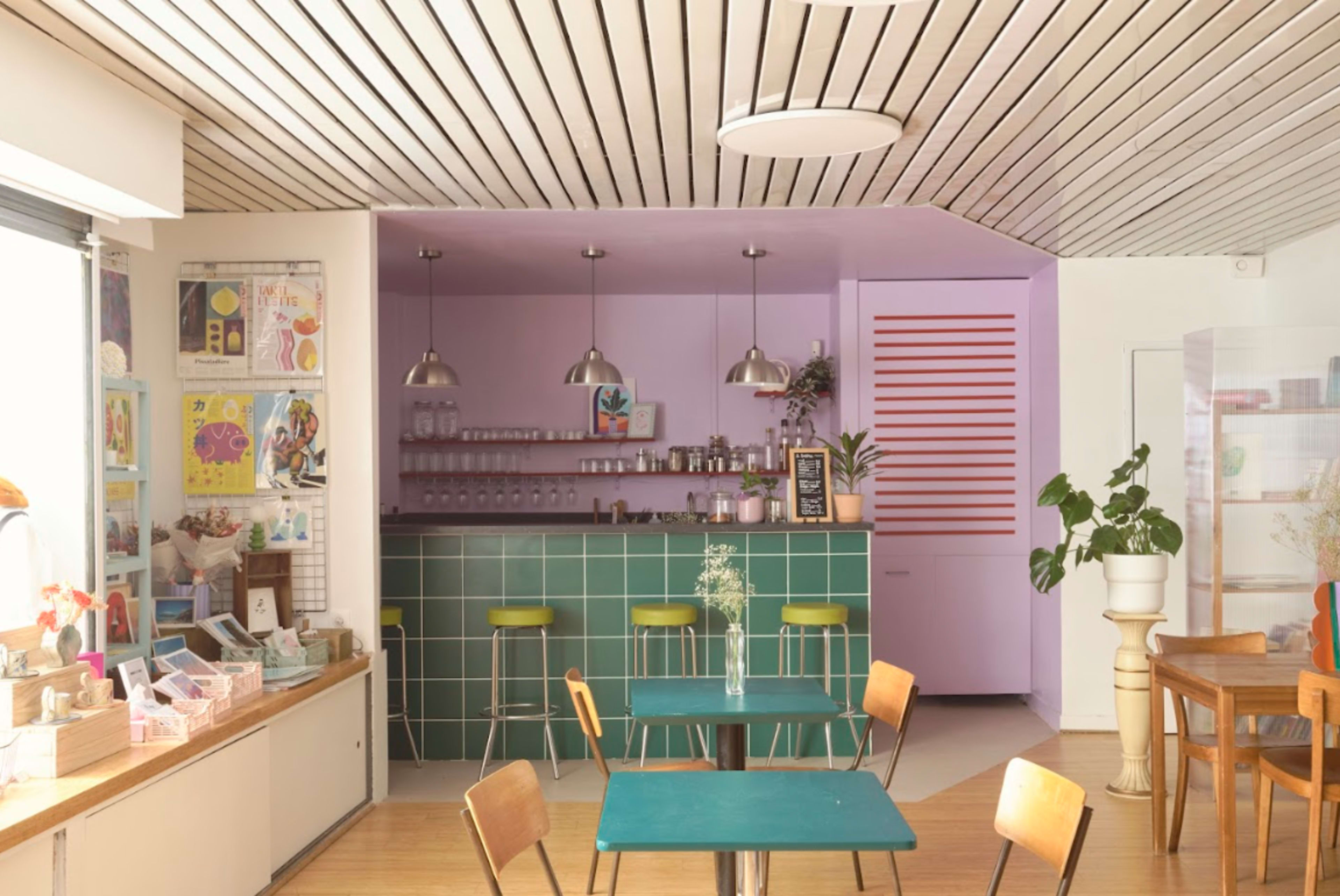 The image shows a modern café interior with a green tile counter, colorful walls, and wooden tables and chairs.