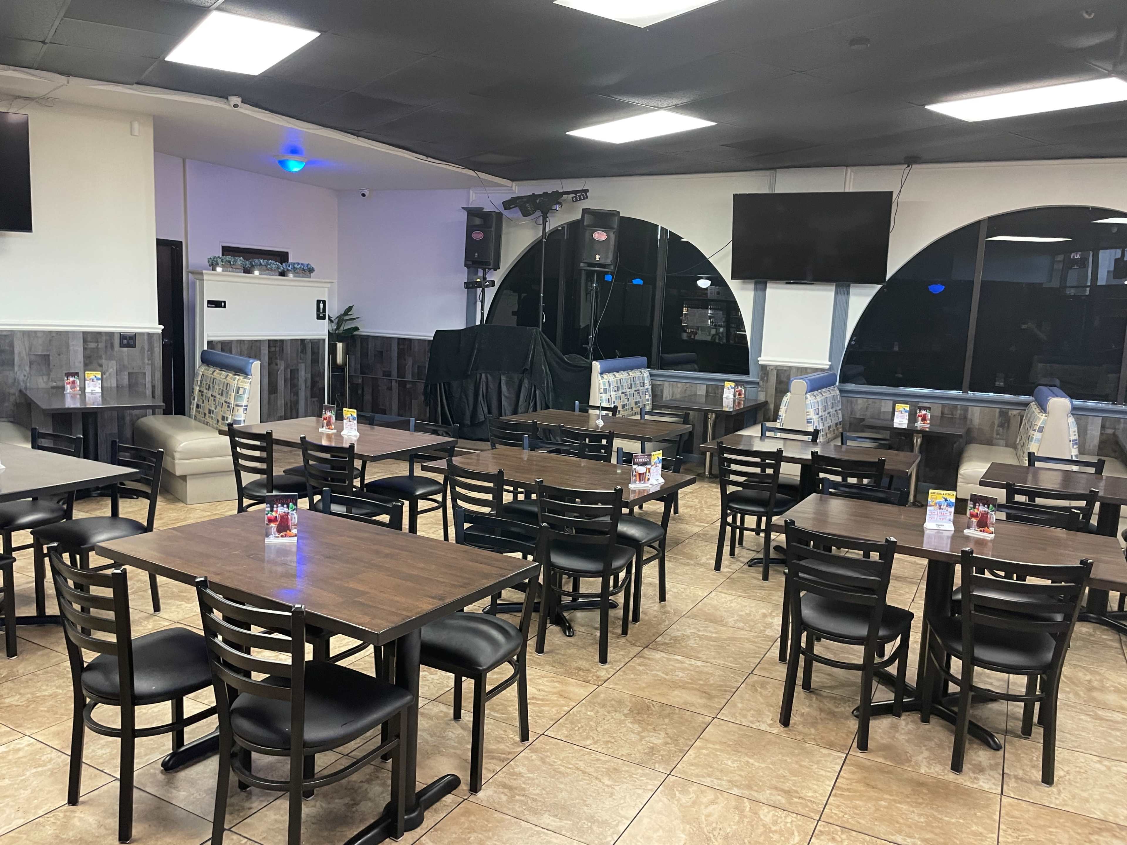 A restaurant interior with multiple tables and chairs arranged in a spacious layout, featuring a stage area and several screens on the walls.