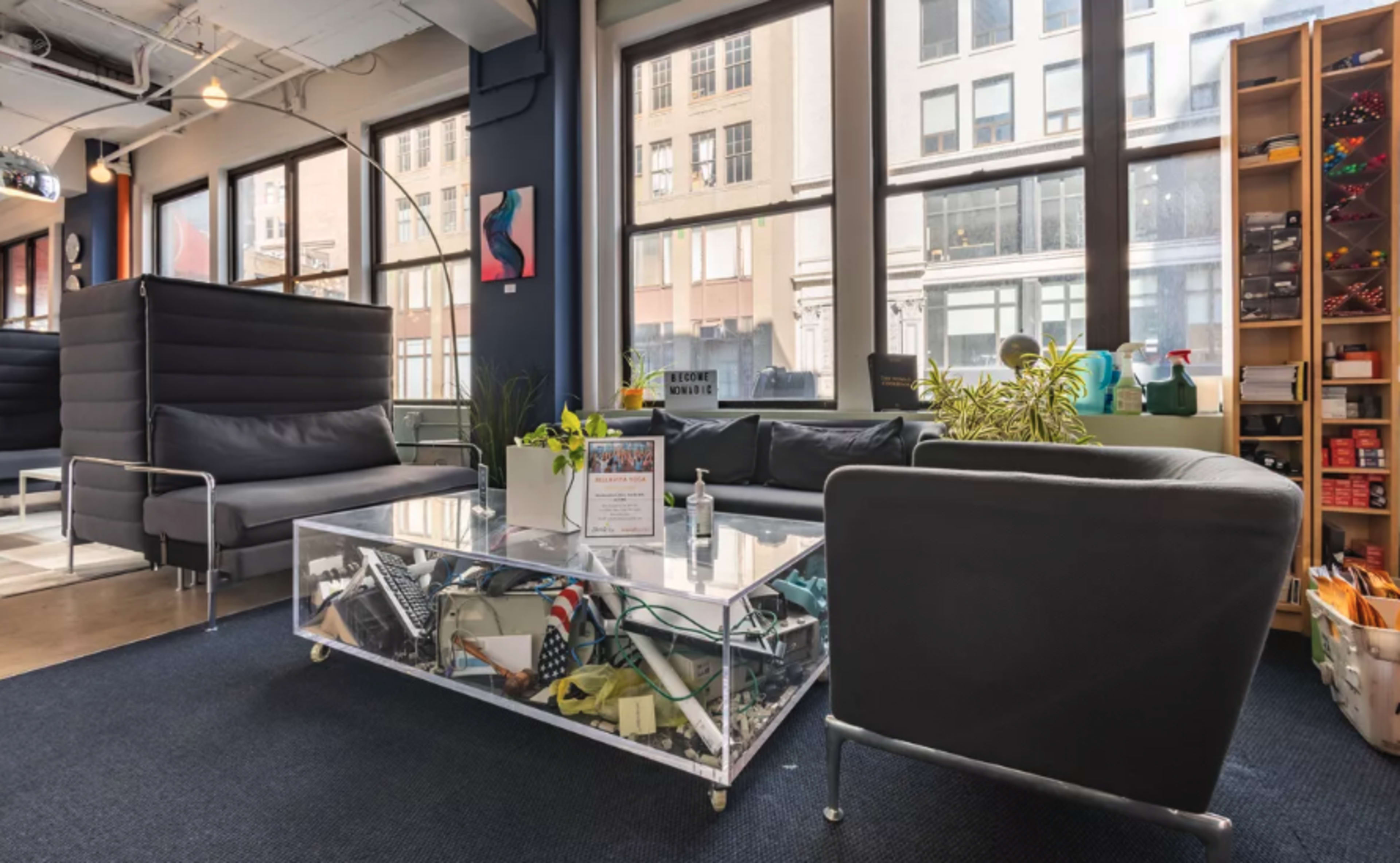 The image shows a cozy seating area with black couches and a glass coffee table surrounded by large windows, revealing a city view.
