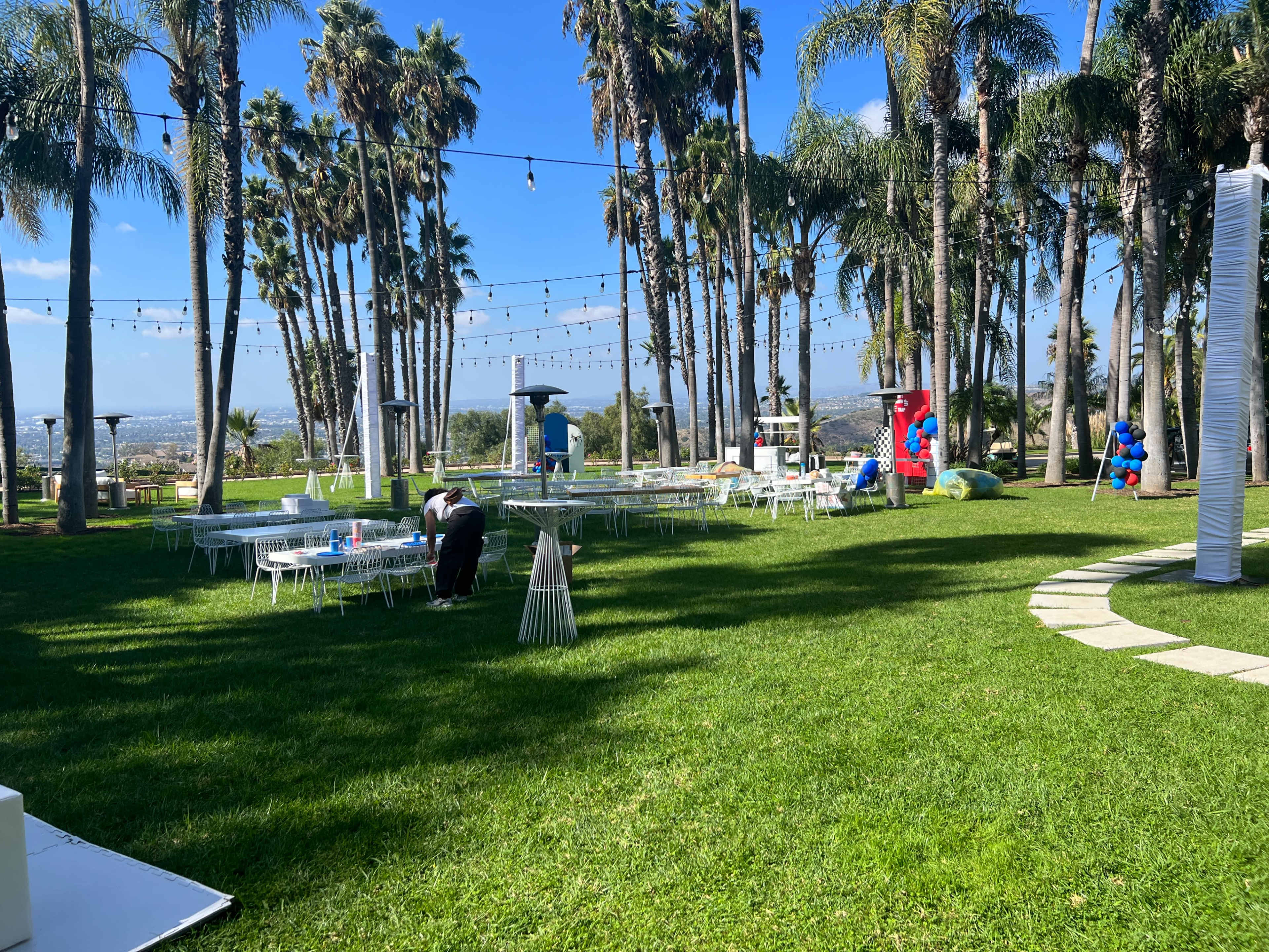 The scene shows a spacious outdoor event area with white tables and chairs arranged on a grass lawn, surrounded by tall palm trees and a clear blue sky.